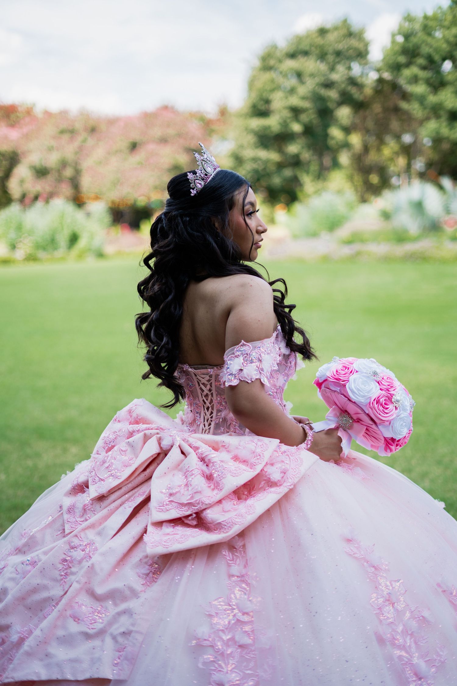 Young woman in pink quinceañera ball gown with tiara and floral bouquet poses in a lush garden.