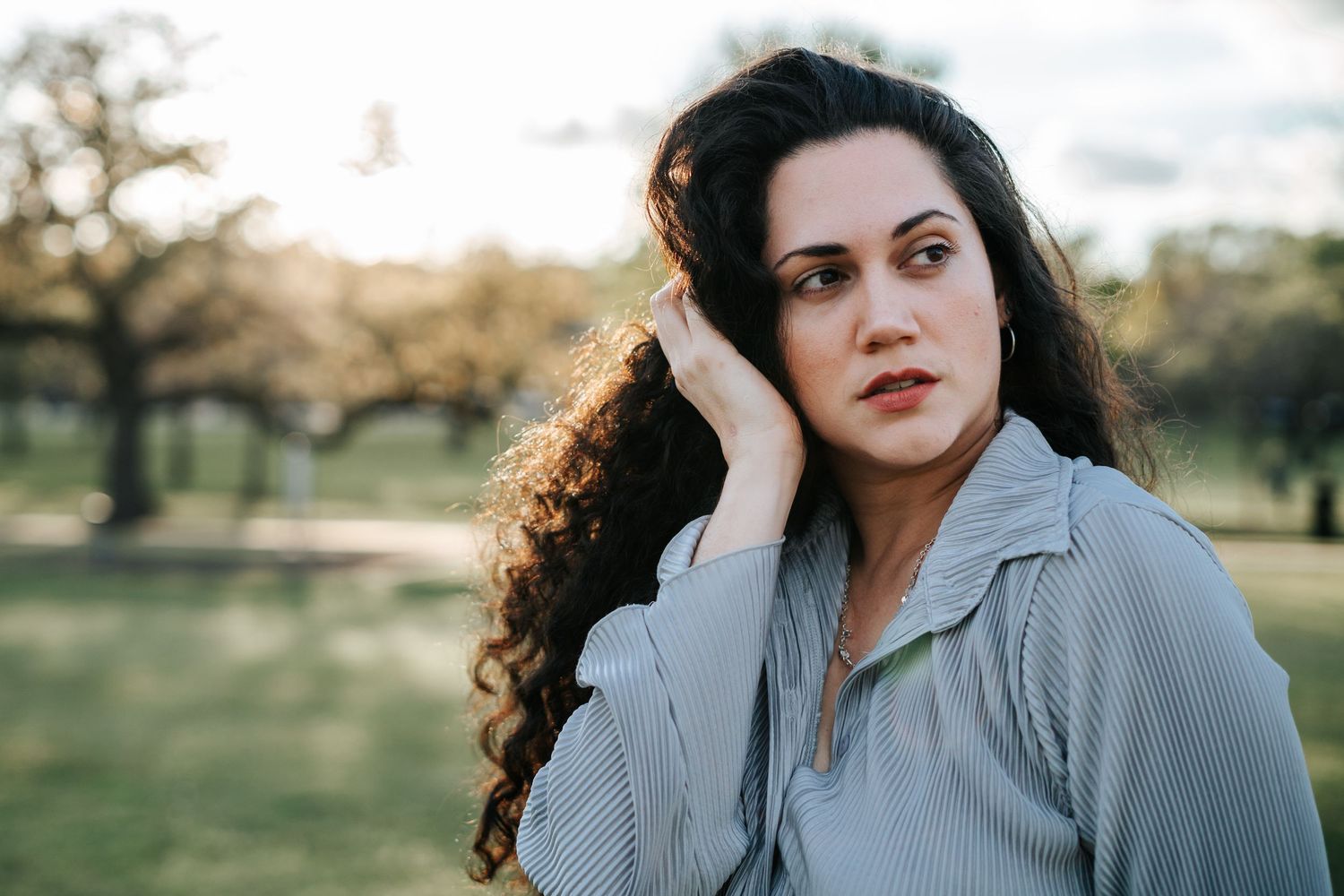 A young woman with curly dark hair poses outdoors in a park, resting her hand near her face in soft golden light.