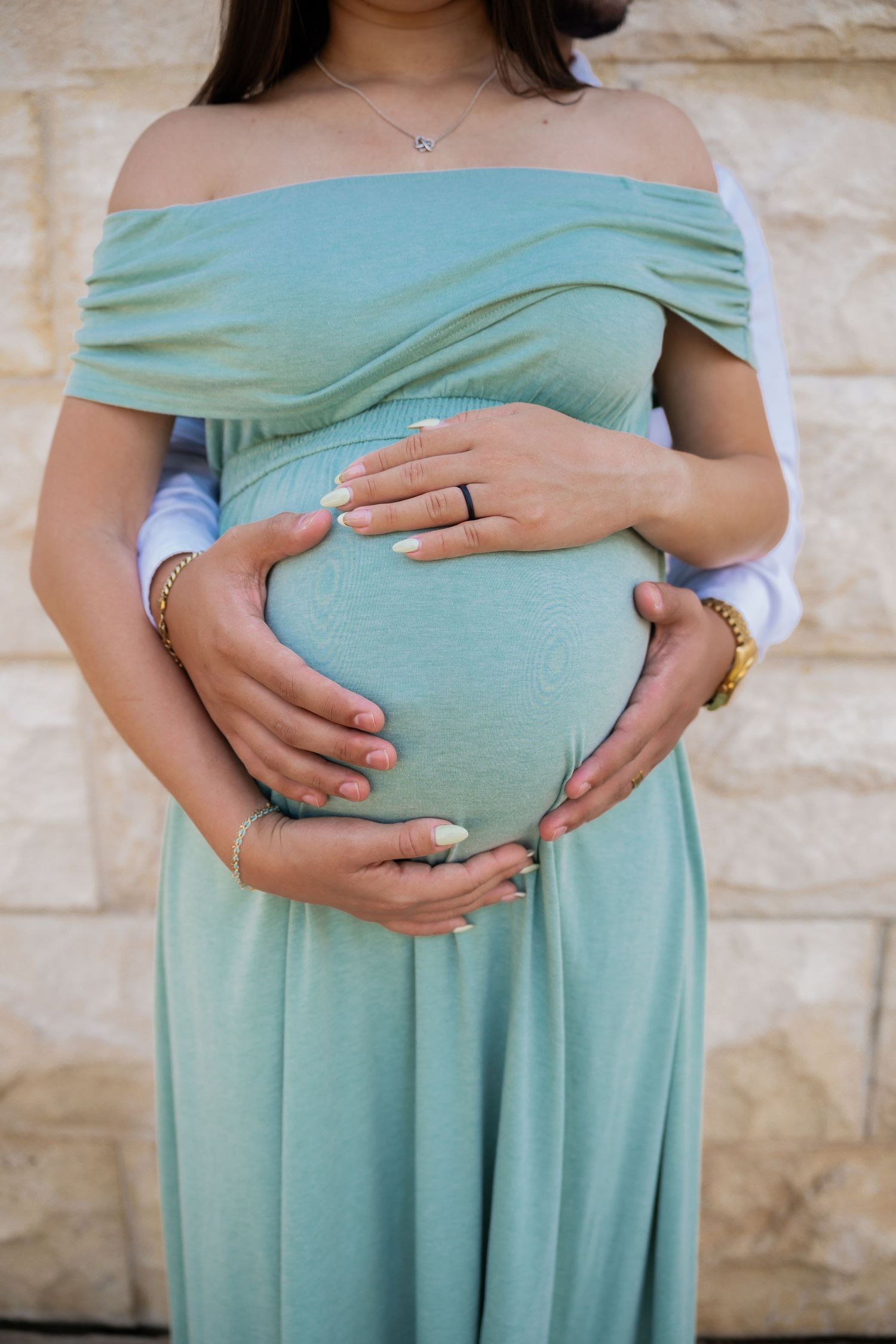 Pregnant woman in sage green off-shoulder dress cradling baby bump with partner's hands, wearing ring and bracelets.