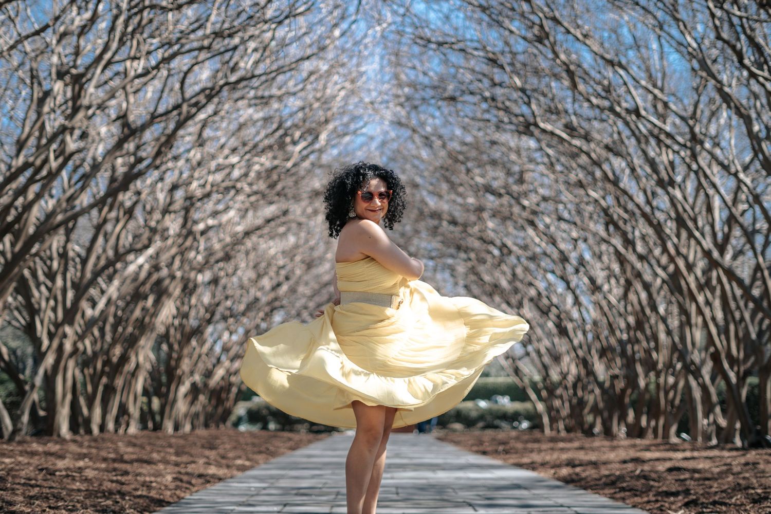 Woman in yellow dress twirling on a tree-lined path with bare branches forming a canopy overhead.