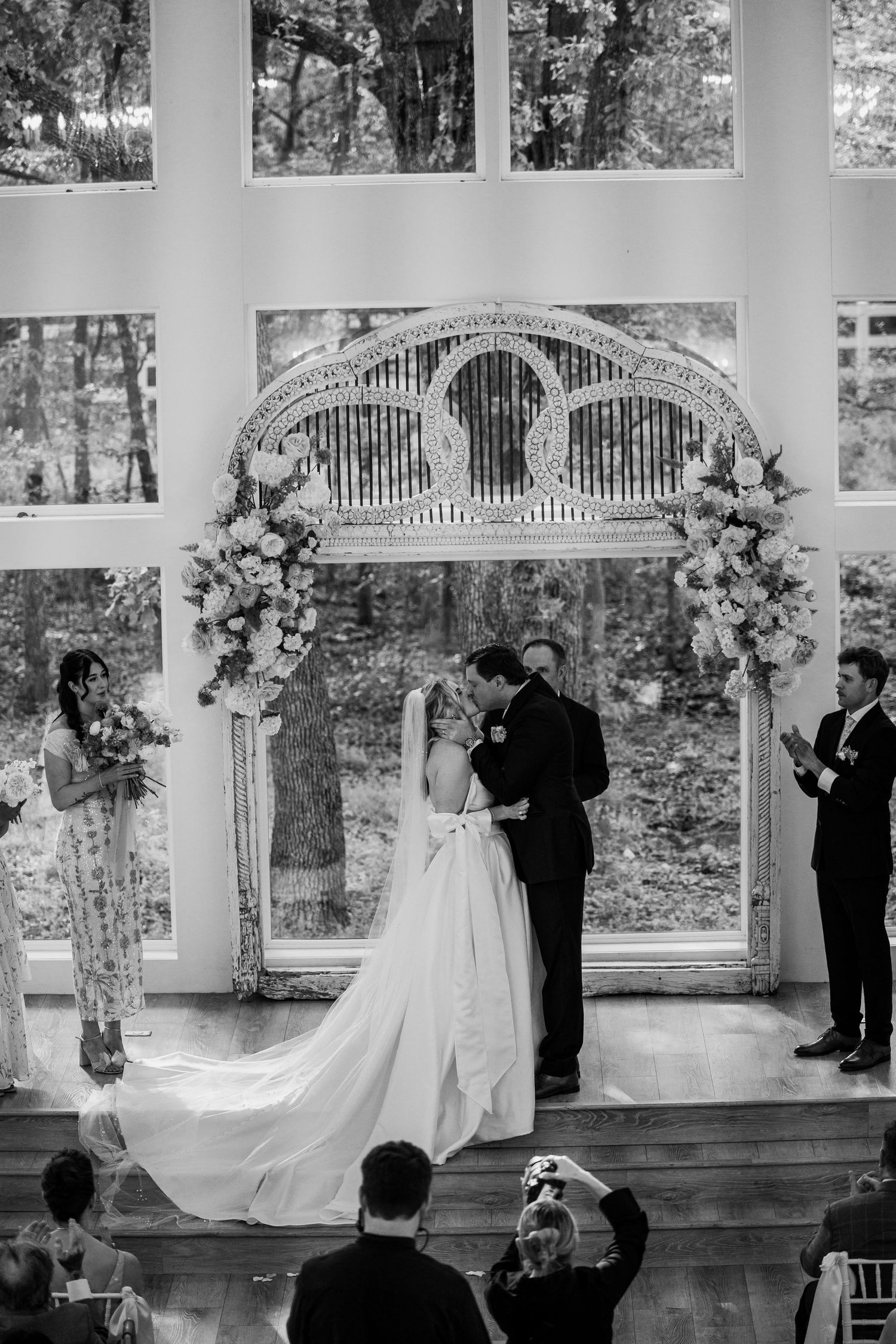 Black and white photo of bride and groom kissing at altar during wedding ceremony with floral arch and large windows.