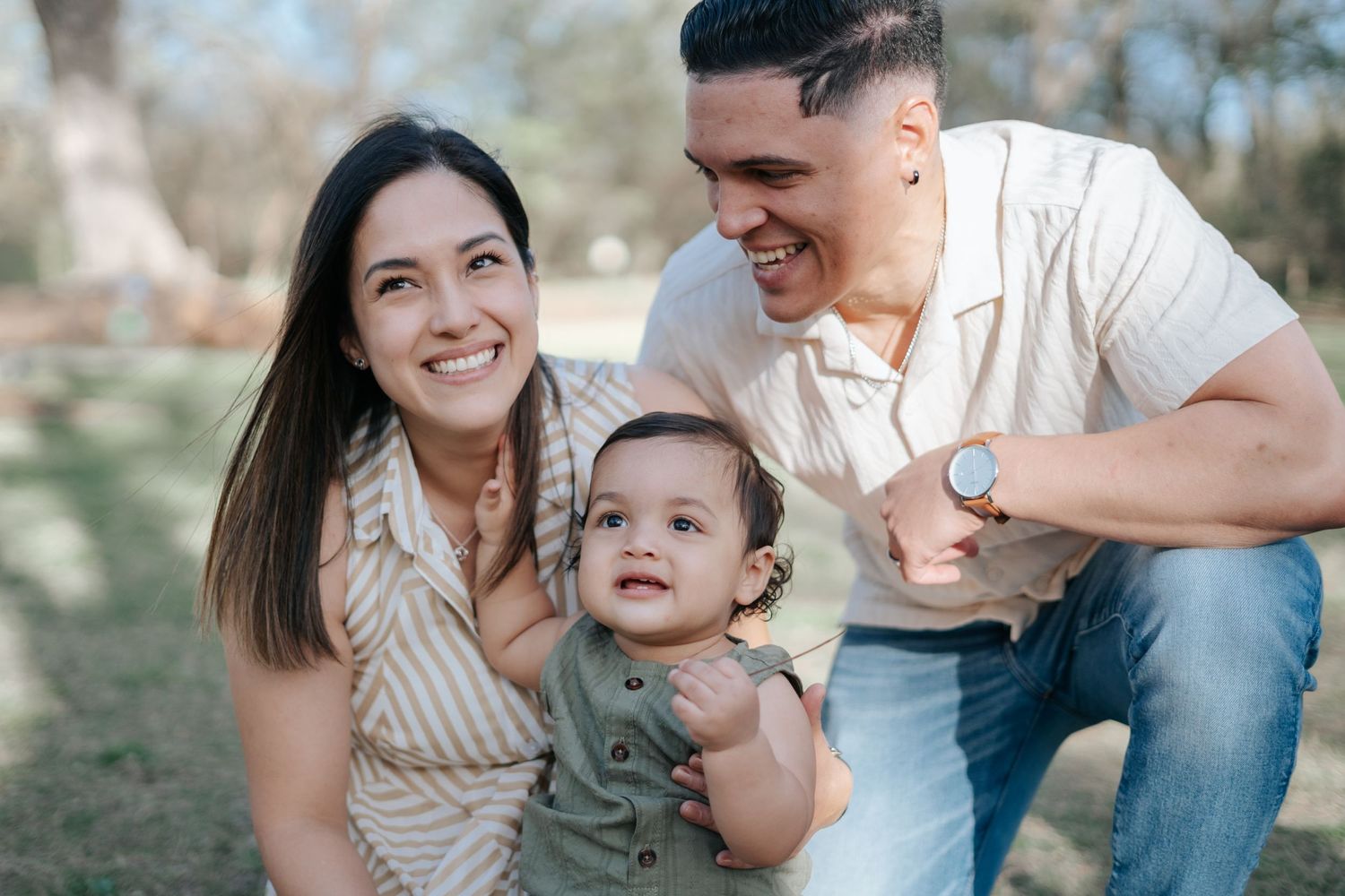 Happy young family with smiling parents and baby enjoying a sunny day outdoors in a park setting.