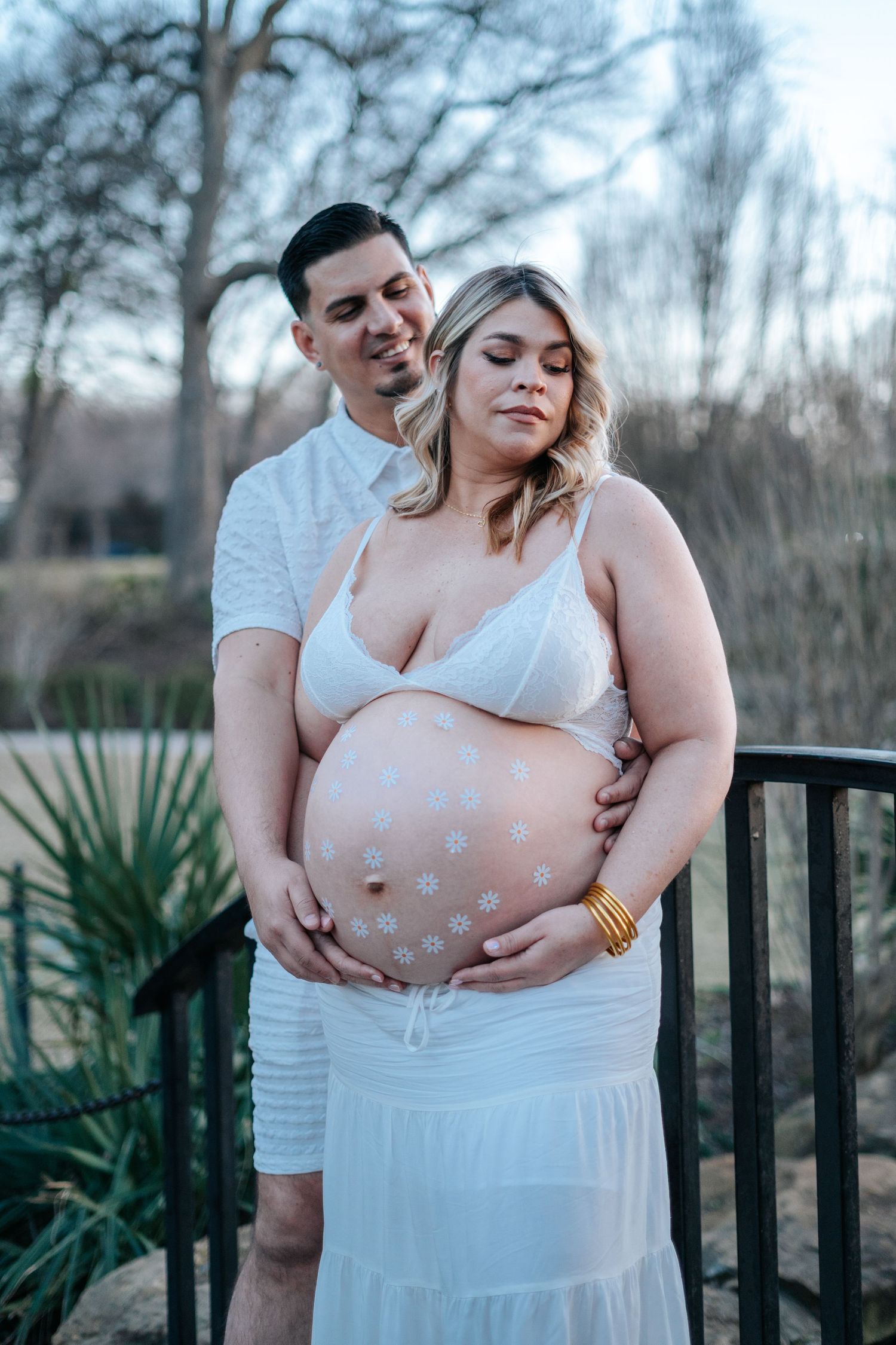 Pregnant couple posing outdoors on a bridge, woman in white maternity outfit with floral belly paint, man embracing her from behind.