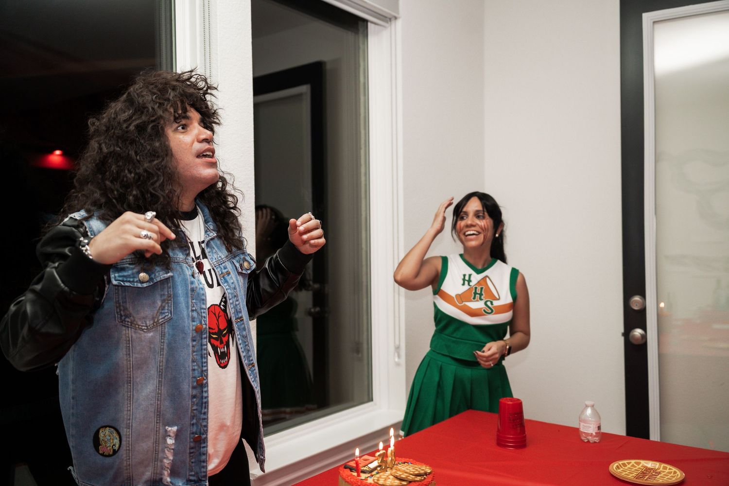 Two people at a party, one in a rock outfit and one in a green cheerleader costume, standing near a red cup on a table.