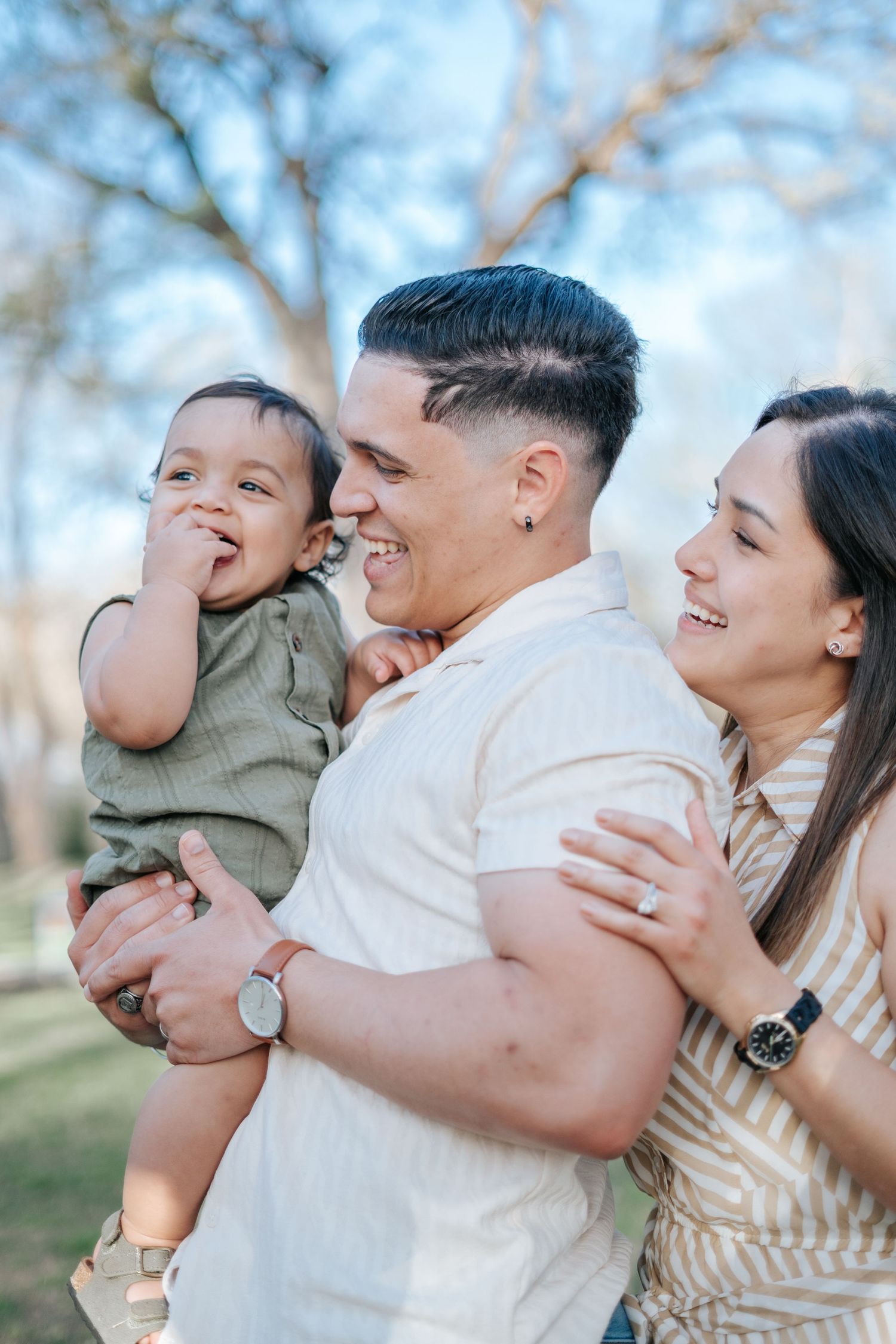 Happy young family with baby boy laughing outdoors in a sunny park setting.