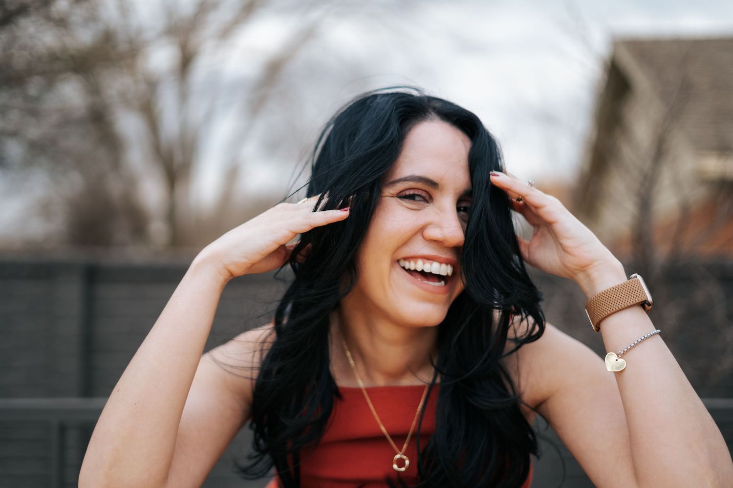 Smiling woman with black hair laughing joyfully, hands in hair, wearing red top and gold necklace outdoors.
