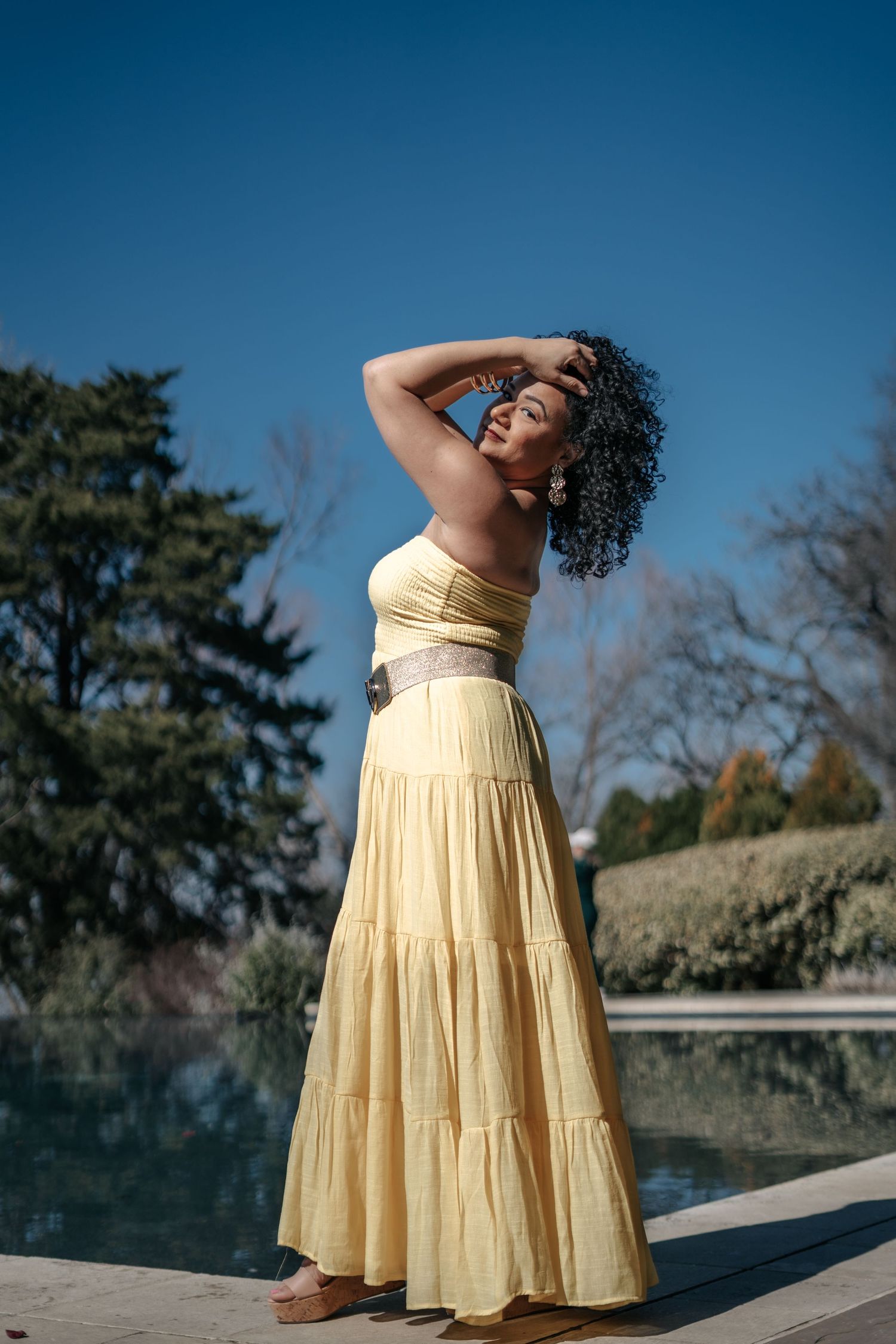 Woman in yellow maxi dress posing outdoors near a reflective pool on a sunny day with blue sky.
