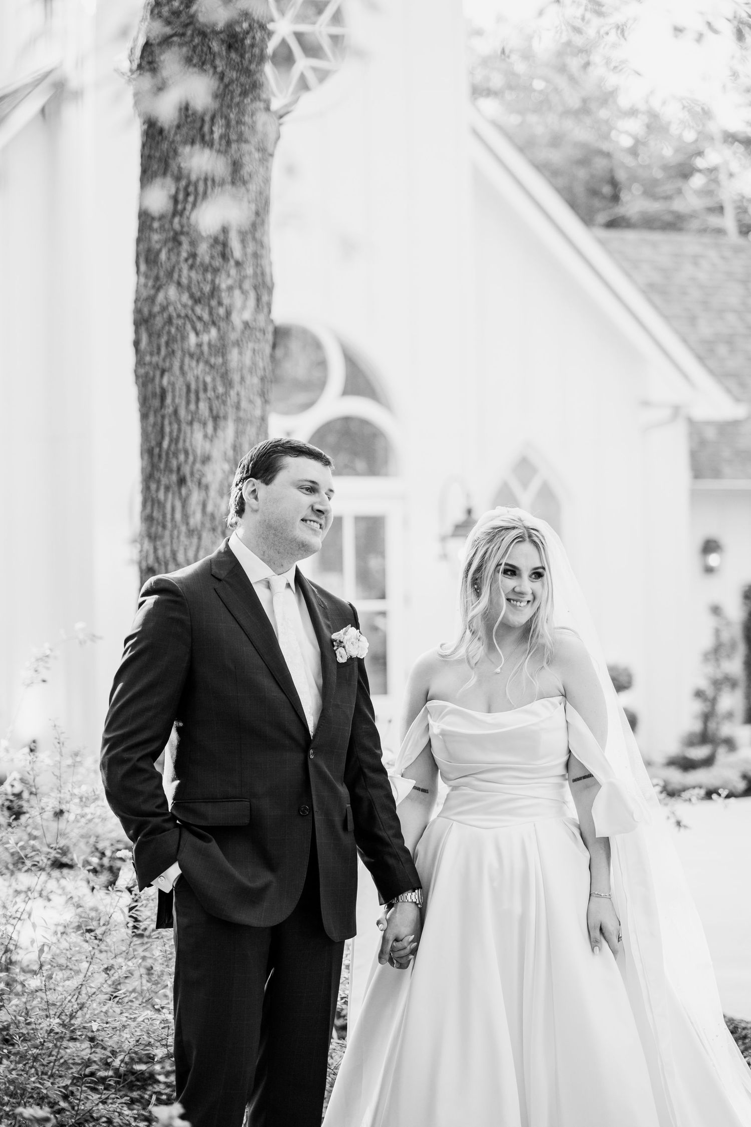 Black and white photo of a smiling bride and groom holding hands outside a white chapel on their wedding day.