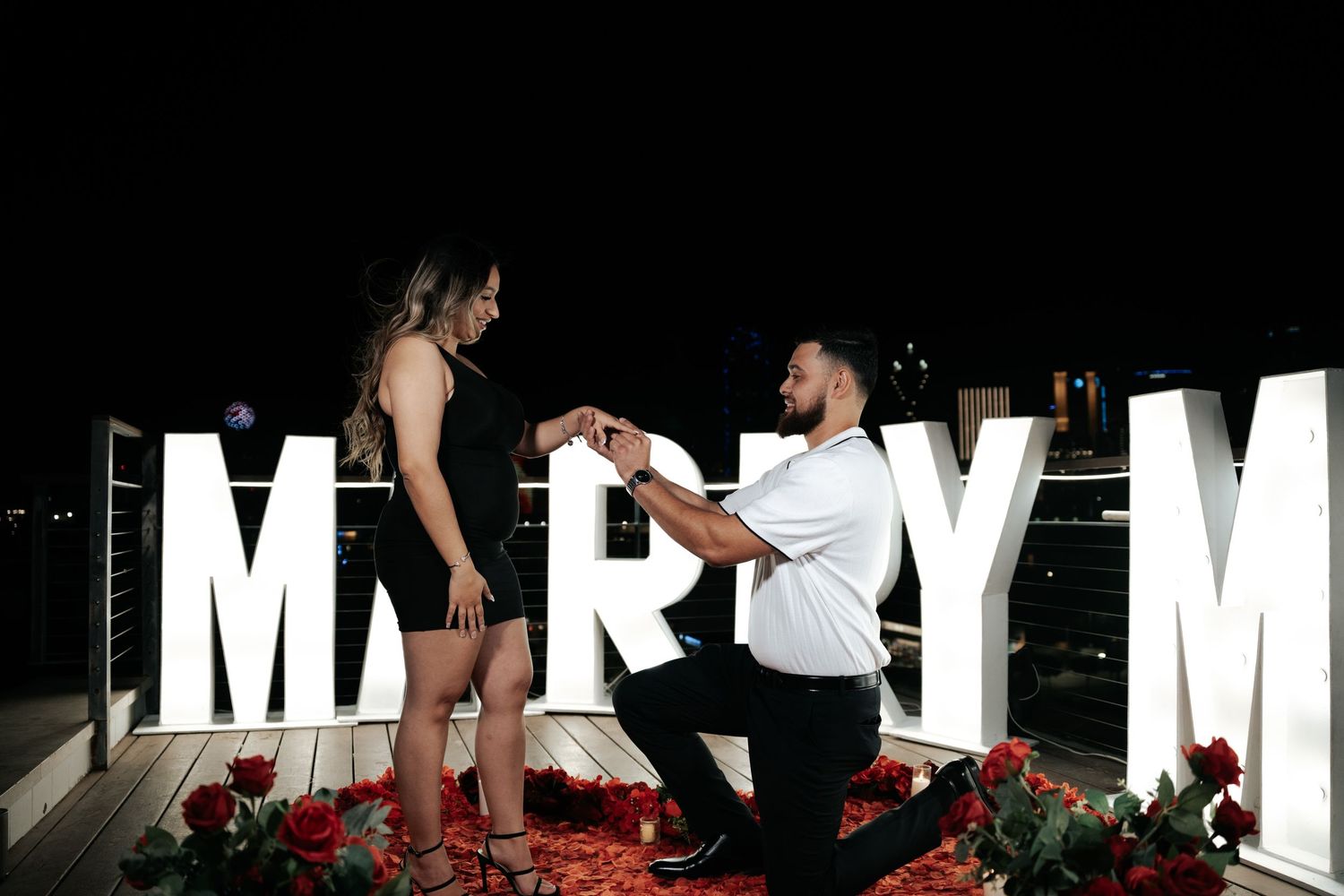 Man proposes to woman on rooftop surrounded by large MARRY ME letters and red roses at night.