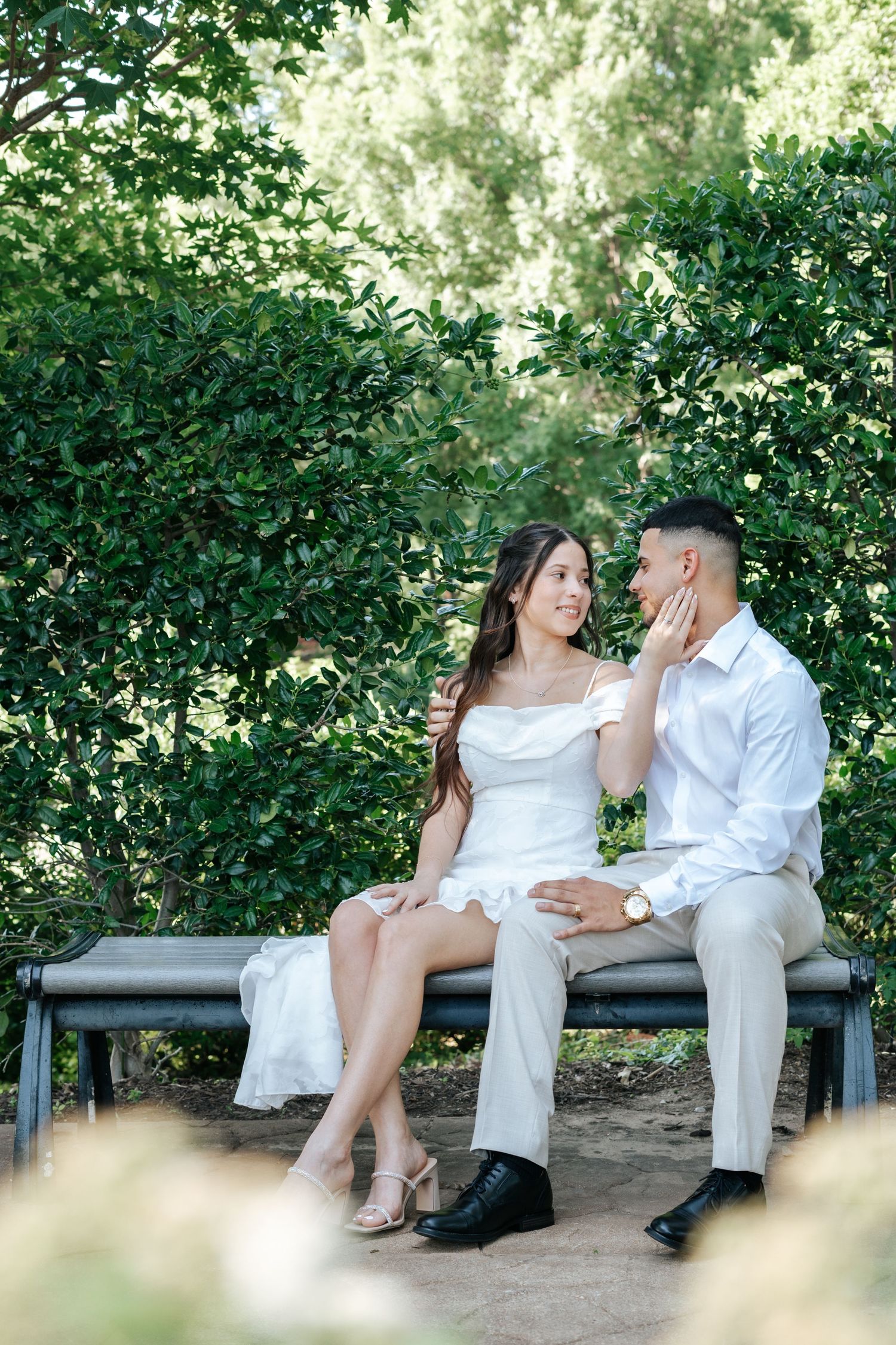 Couple in white outfits sitting on a bench surrounded by lush green hedges in a romantic outdoor setting.