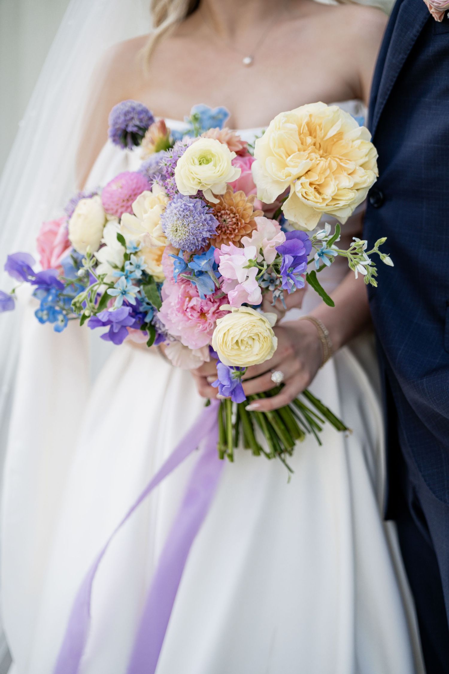 Bride holding a colorful wedding bouquet with yellow roses, pink blooms, and purple flowers, tied with a lavender ribbon.