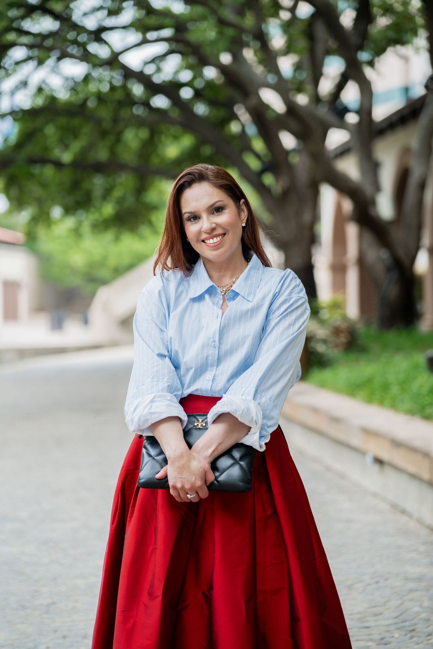 Smiling woman in blue striped blouse and red maxi skirt holding a gray clutch outdoors near a large tree.