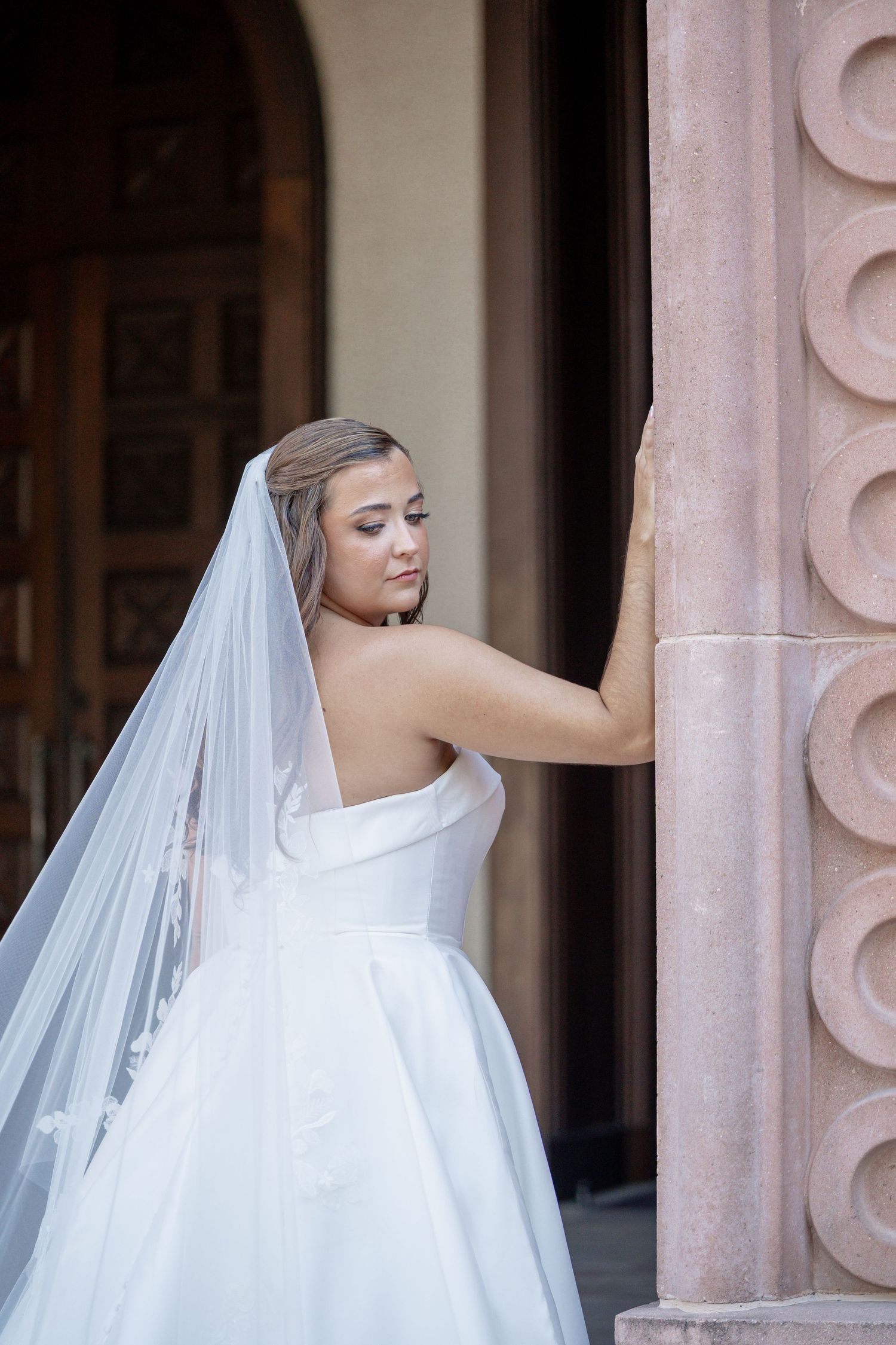 Bride in strapless white ball gown and long veil poses gracefully beside ornate pink stone column at elegant venue.
