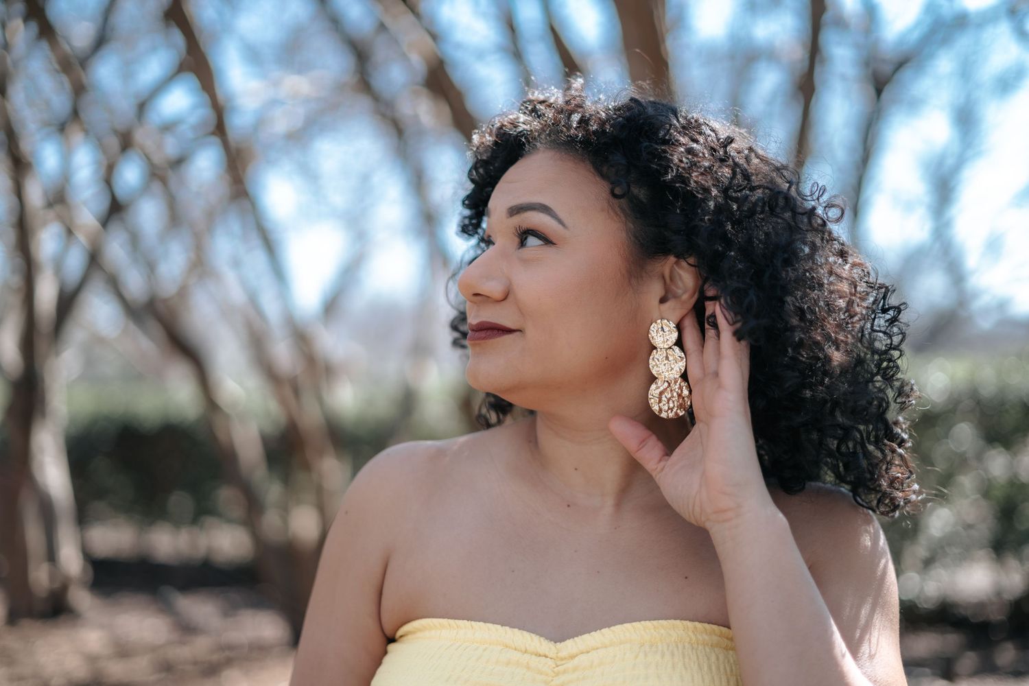 Young woman with curly hair wearing yellow strapless top and gold earrings posing outdoors among trees.