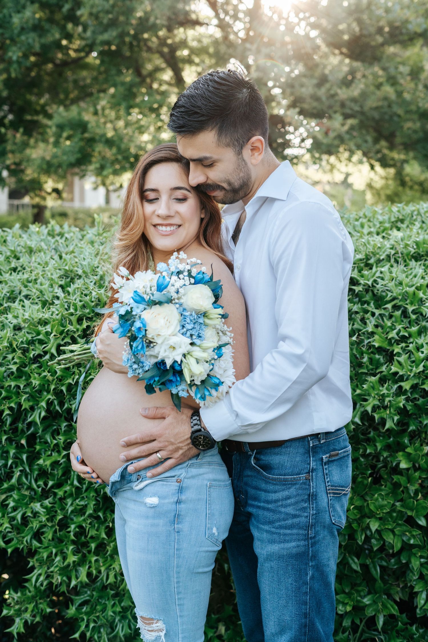 Pregnant couple embracing outdoors, woman holding blue and white flower bouquet, both wearing jeans, green hedges background.