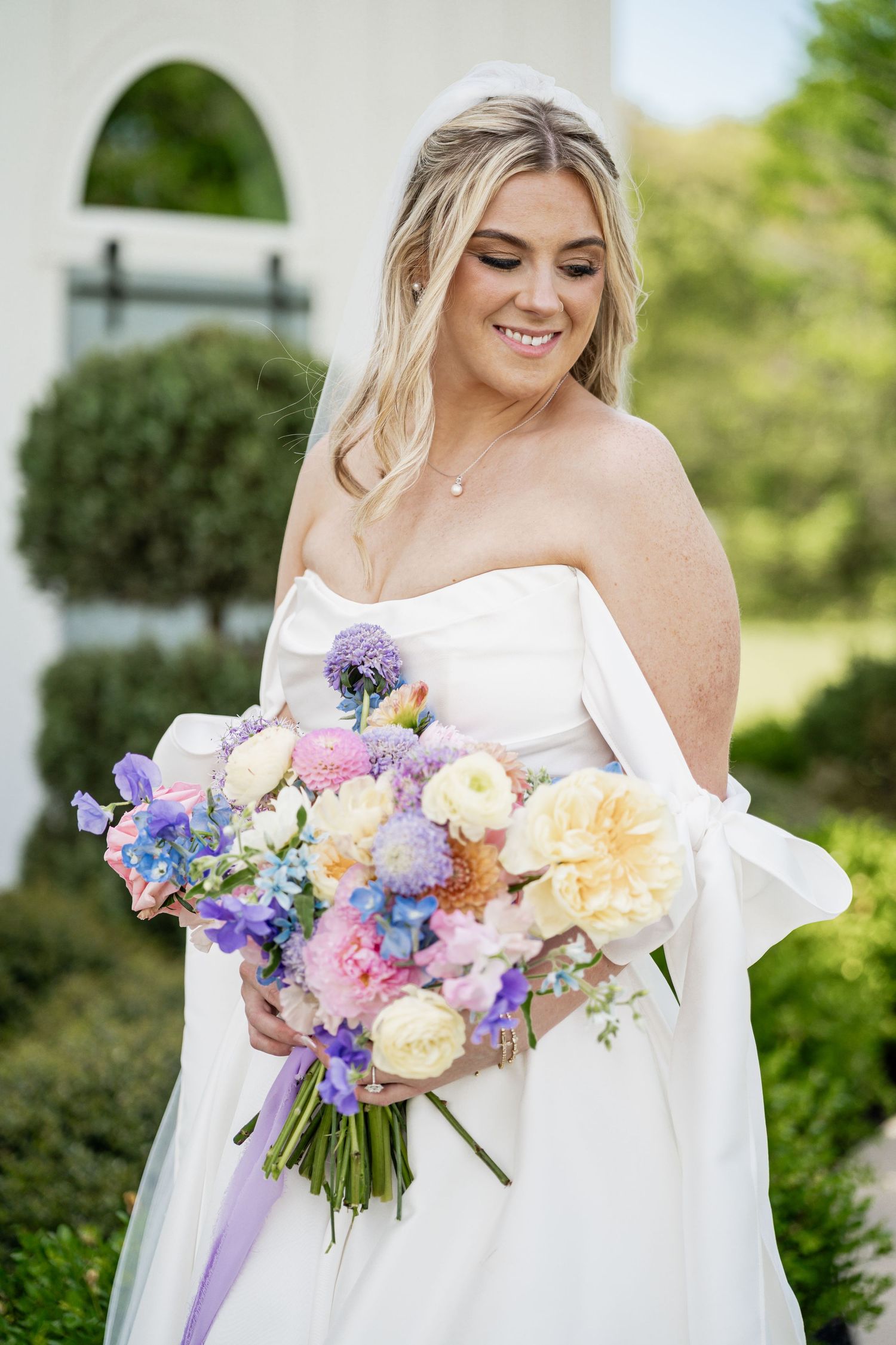 Smiling bride in white off shoulder gown holding colorful spring bouquet with pink, blue, and yellow flowers outdoors.
