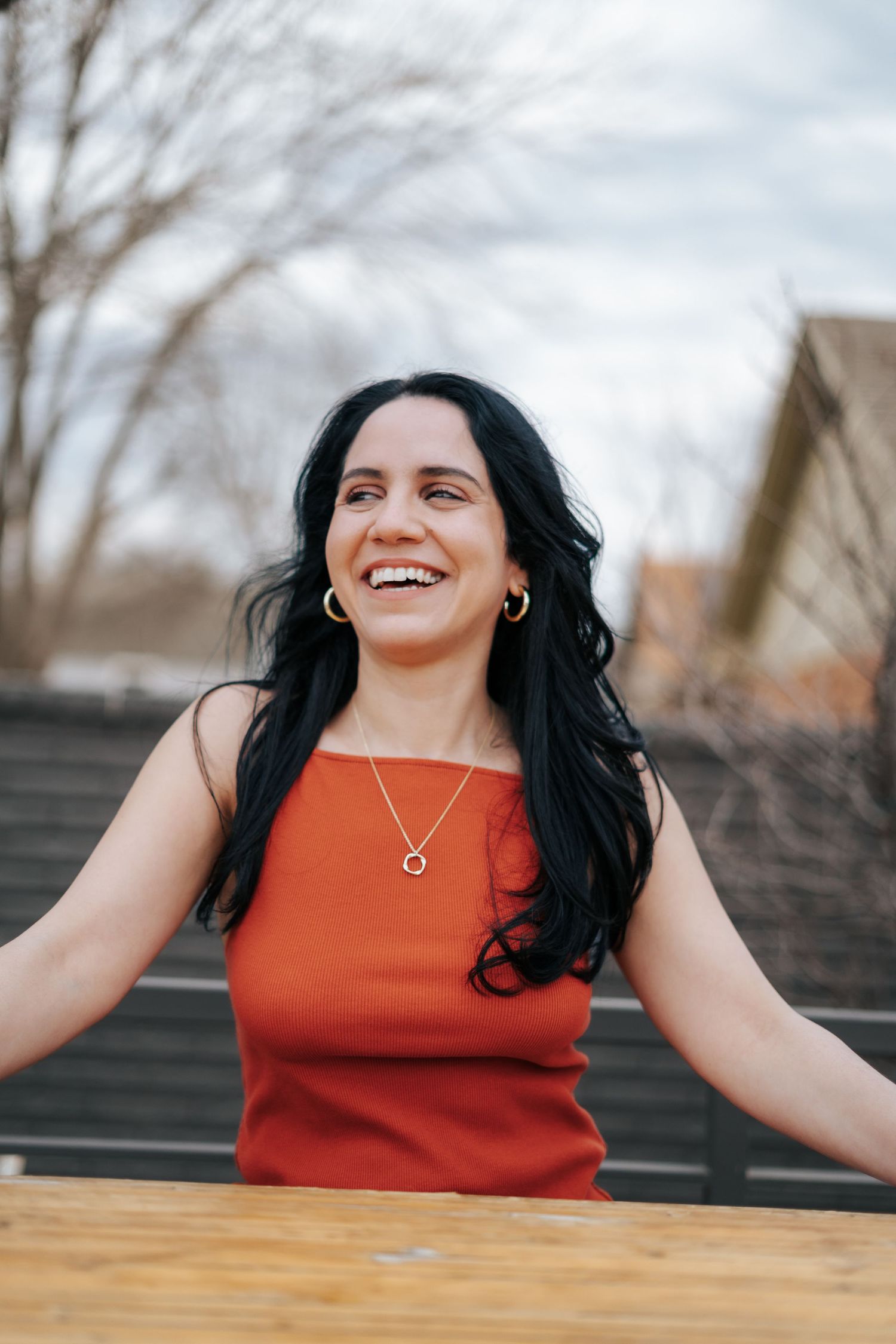Smiling woman with black hair wearing a red dress and gold hoop earrings, laughing outdoors near a wooden table.