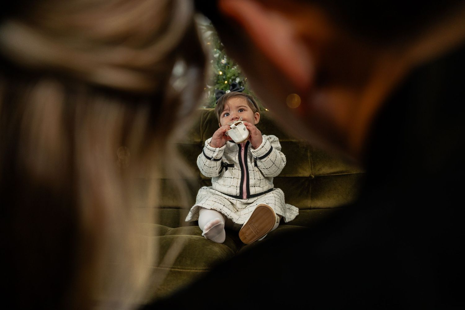 Baby in knit outfit sits on steps drinking from cup, framed by two adults, with Christmas tree in background.