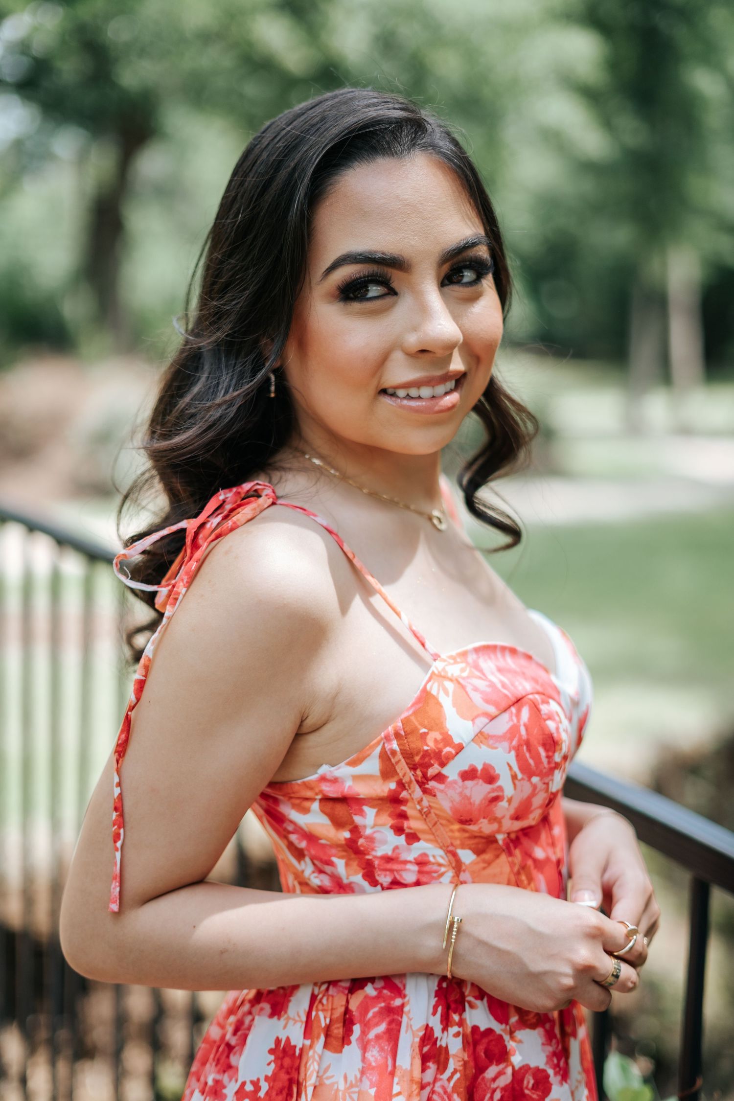 Young woman in red floral dress smiling outdoors near iron railing with lush green trees background.