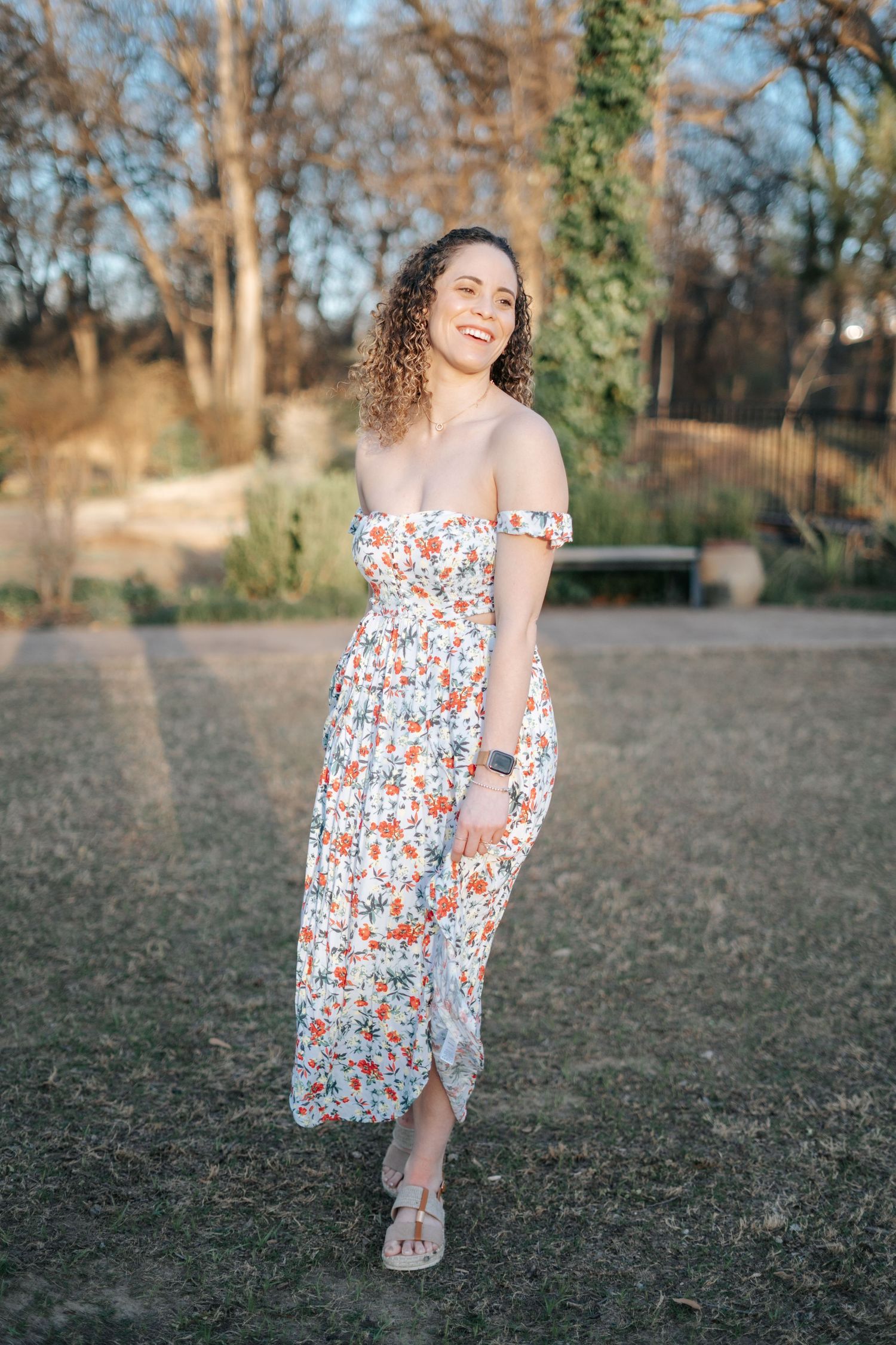 Woman in floral off shoulder midi dress smiling in a sunny park setting with curly hair and sandals.