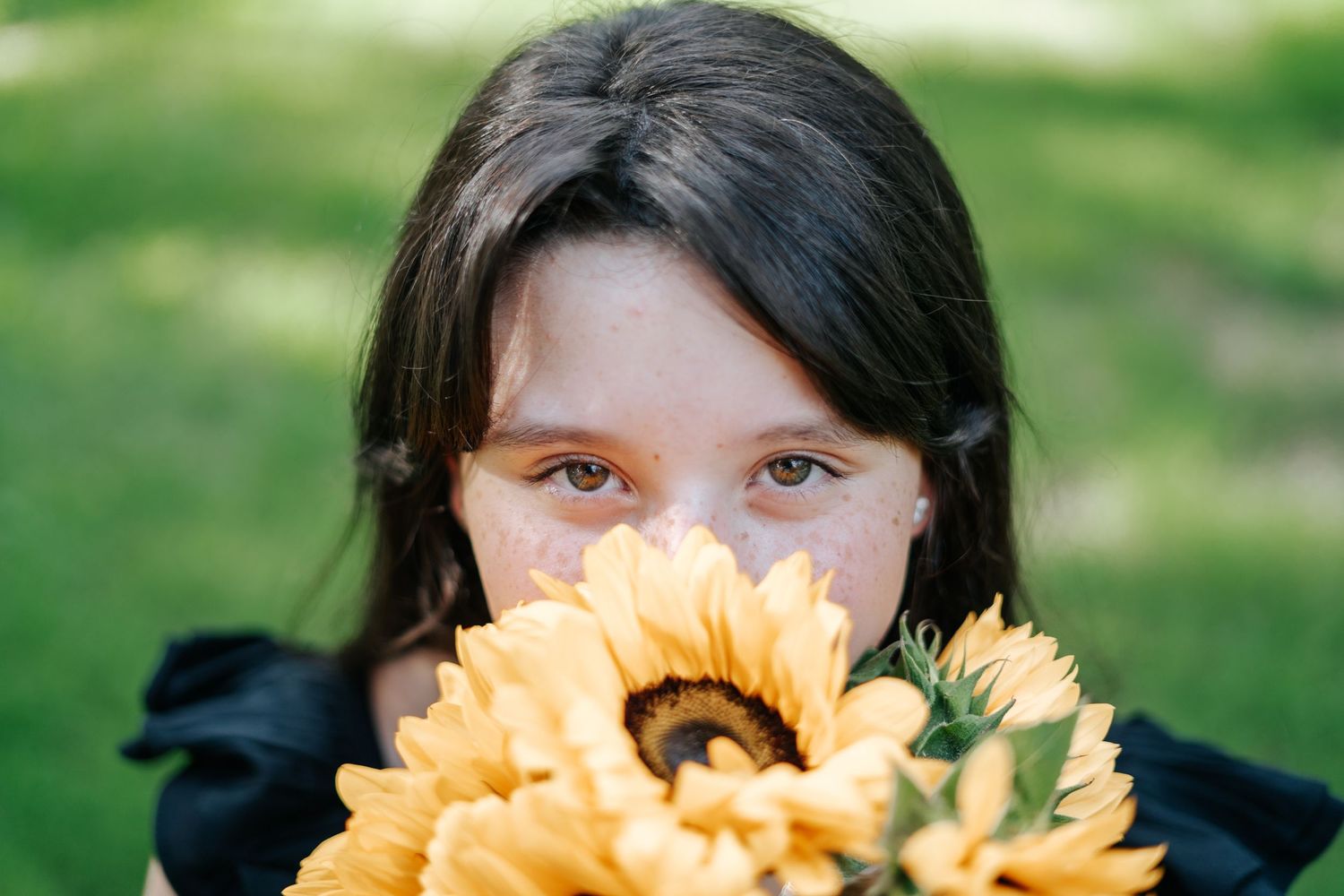 Young girl with dark hair holds a bright sunflower bouquet up to her face outdoors on a green lawn.