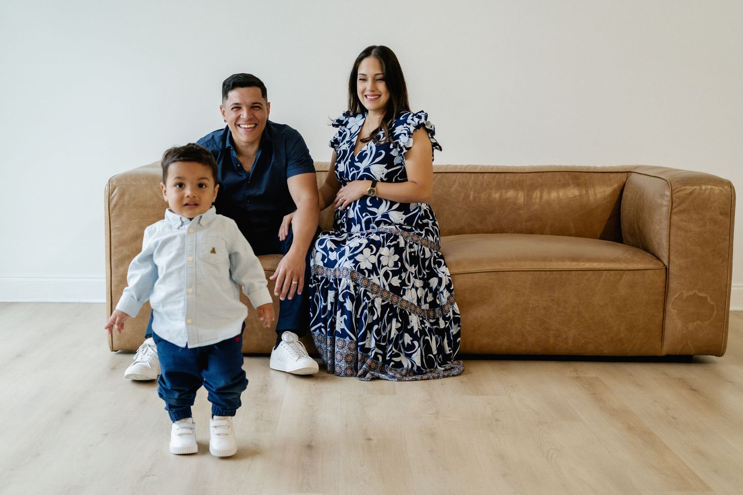 Family portrait of parents sitting on brown leather sofa with toddler boy standing in front on light wood floor.