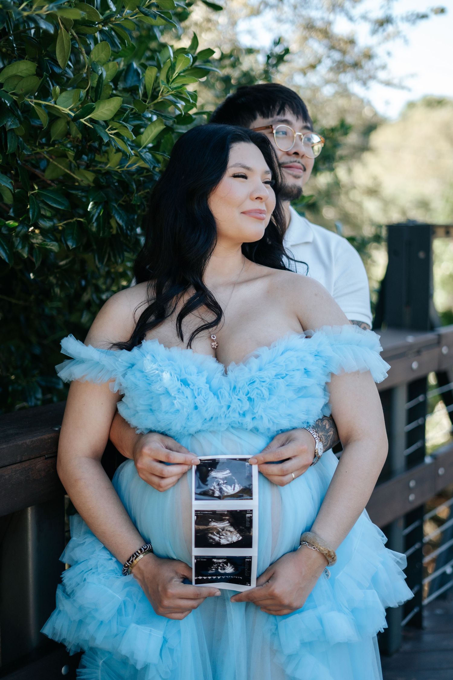 Couple in a park holding ultrasound photos, woman in blue ruffled gown, pregnancy announcement photo shoot.