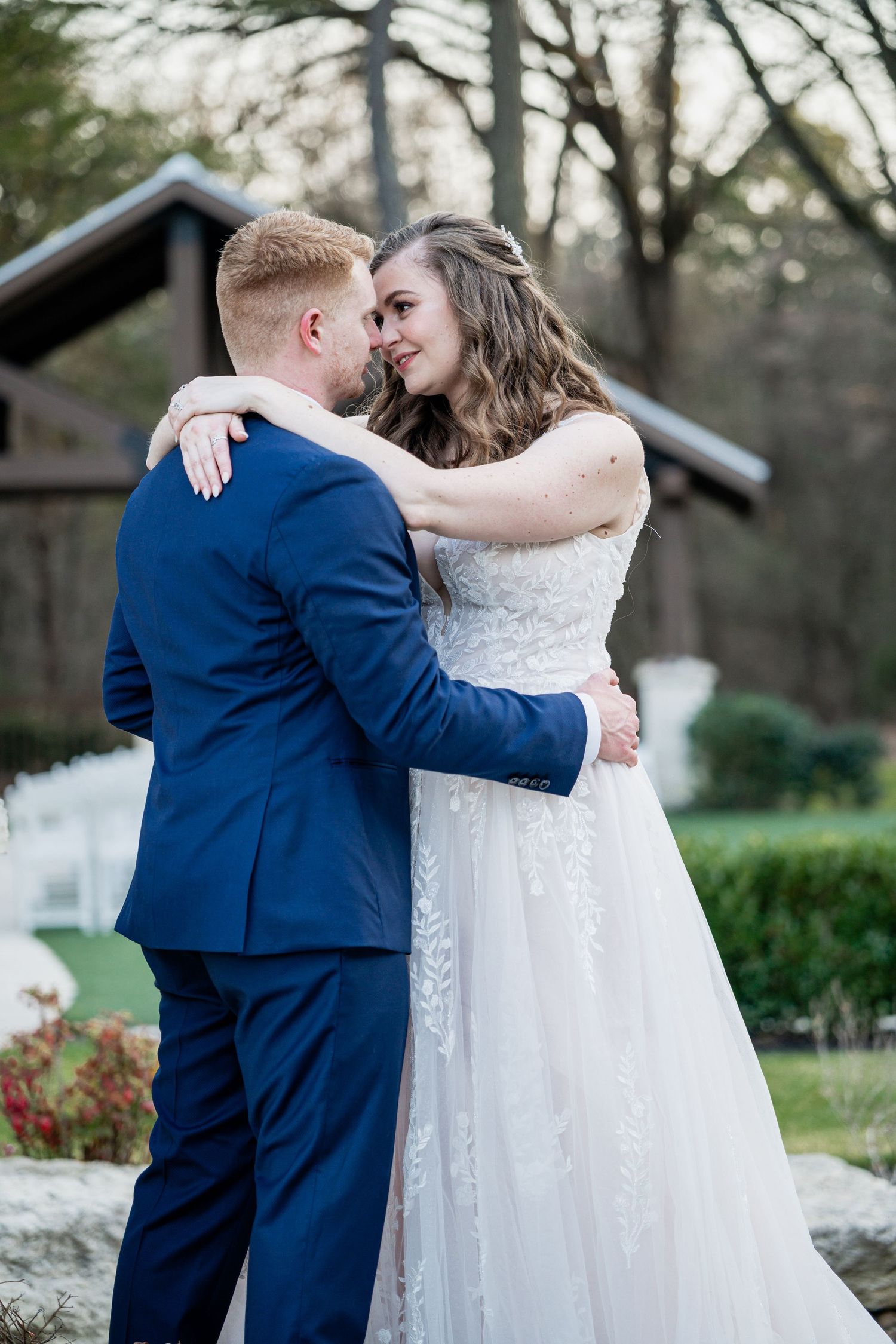 A bride and groom share a romantic embrace outdoors, with the groom in a blue suit and bride in a lace wedding gown.