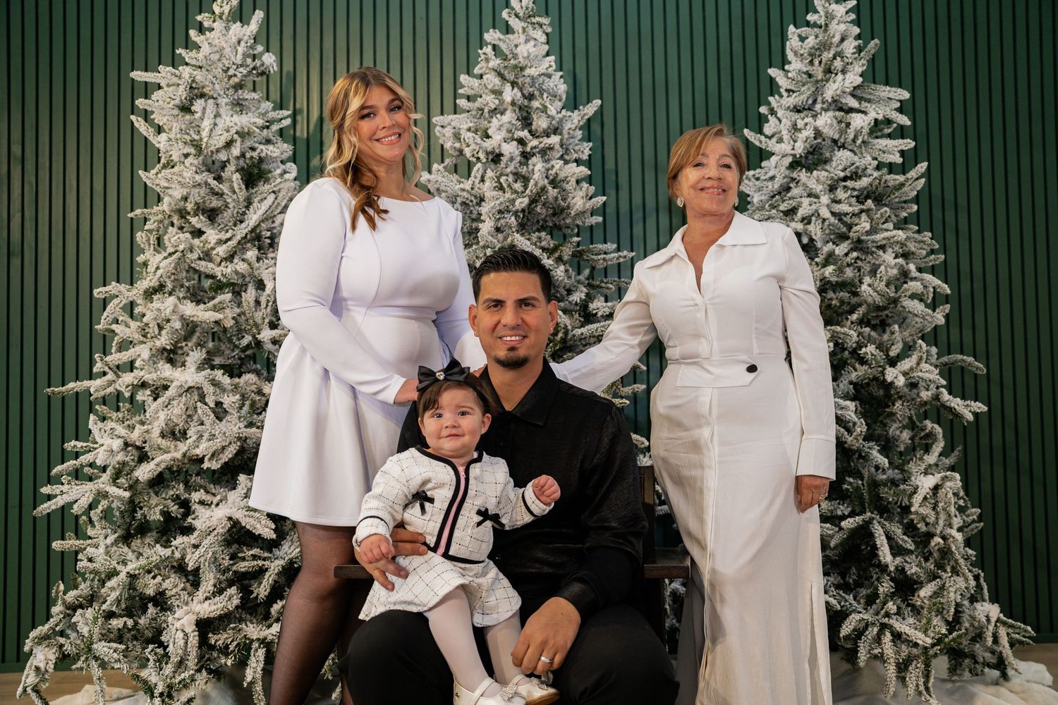 Family of four poses before flocked Christmas trees in a festive holiday portrait with elegant black and white outfits.
