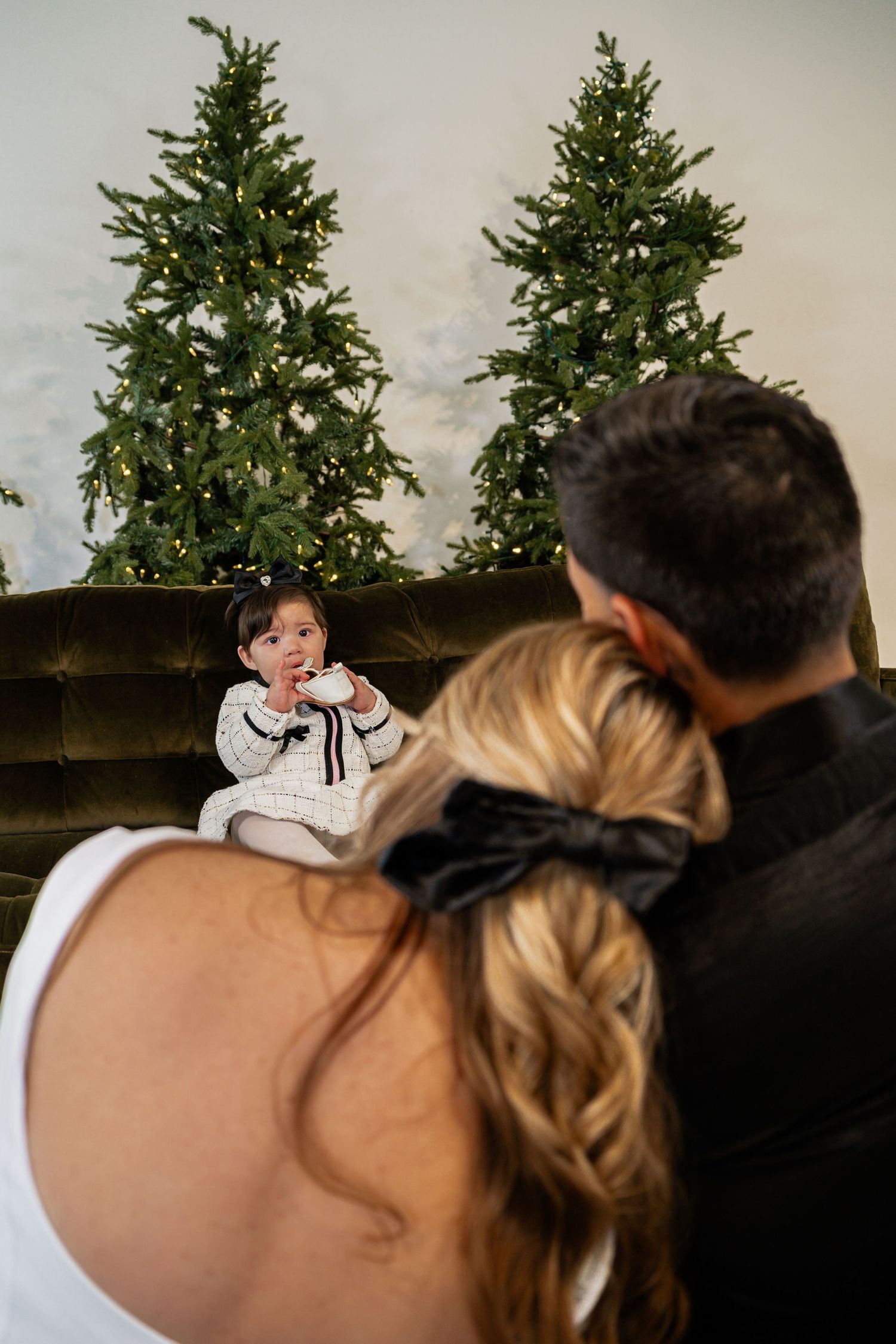 A toddler sits on a green velvet sofa between lit Christmas trees while parents watch from the foreground.