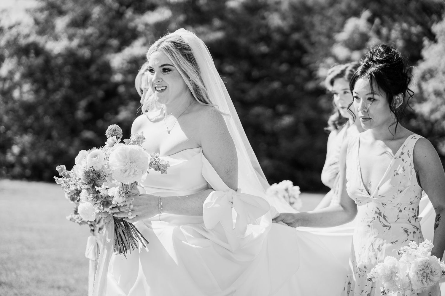 Black and white photo of a smiling bride in a white gown and veil holding a bouquet, flanked by two bridesmaids outdoors.