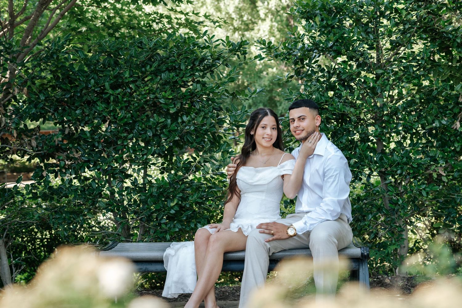 Couple sitting on a park bench surrounded by lush green bushes, woman in white dress, man in white shirt.