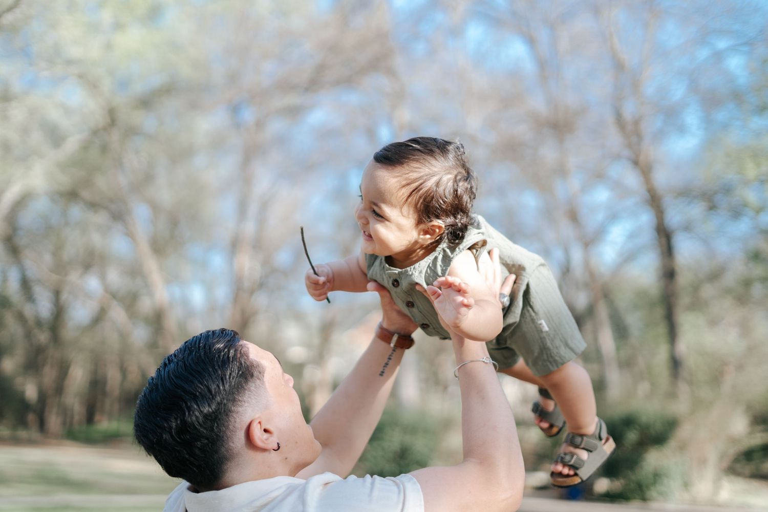 A father joyfully lifts his smiling baby into the air outdoors in a sunny park surrounded by trees.