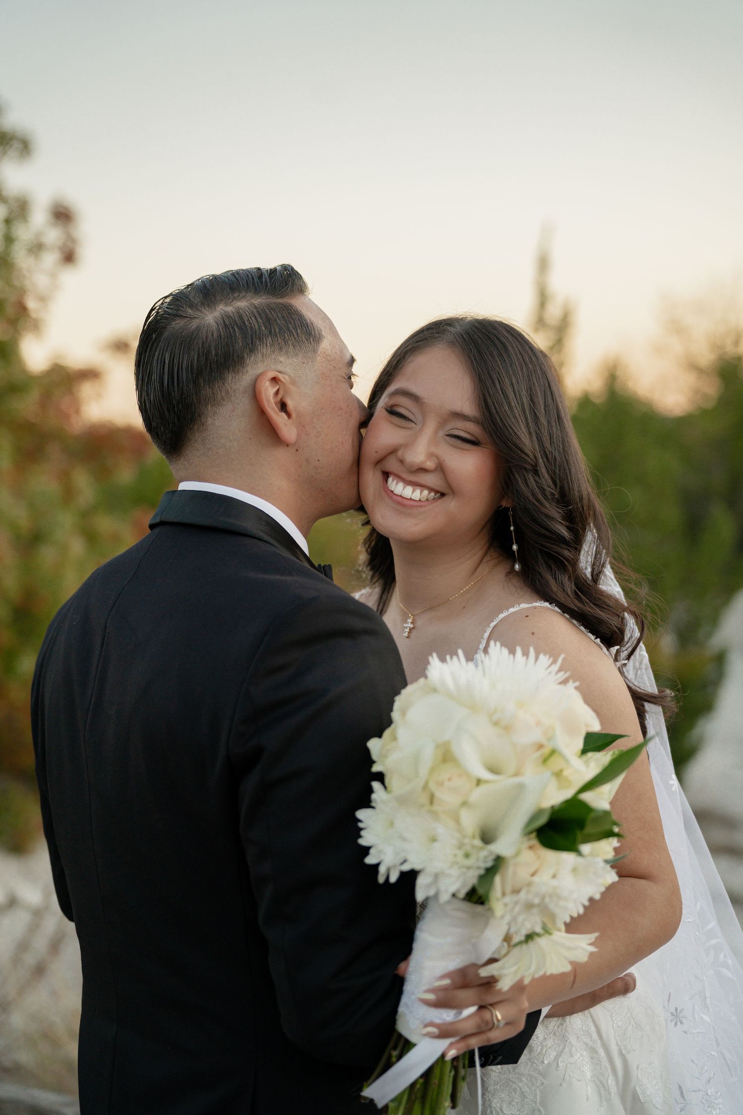 Groom in black suit kissing bride on cheek as she smiles holding white floral bouquet at outdoor wedding.