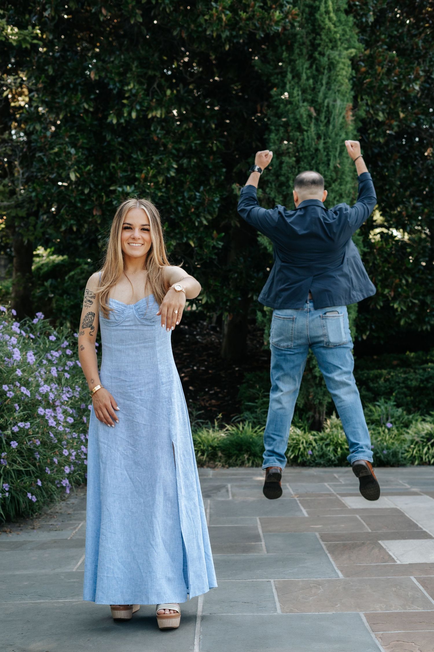 Woman in blue maxi dress smiles outdoors as man in jeans jumps with raised fists in celebration behind her.