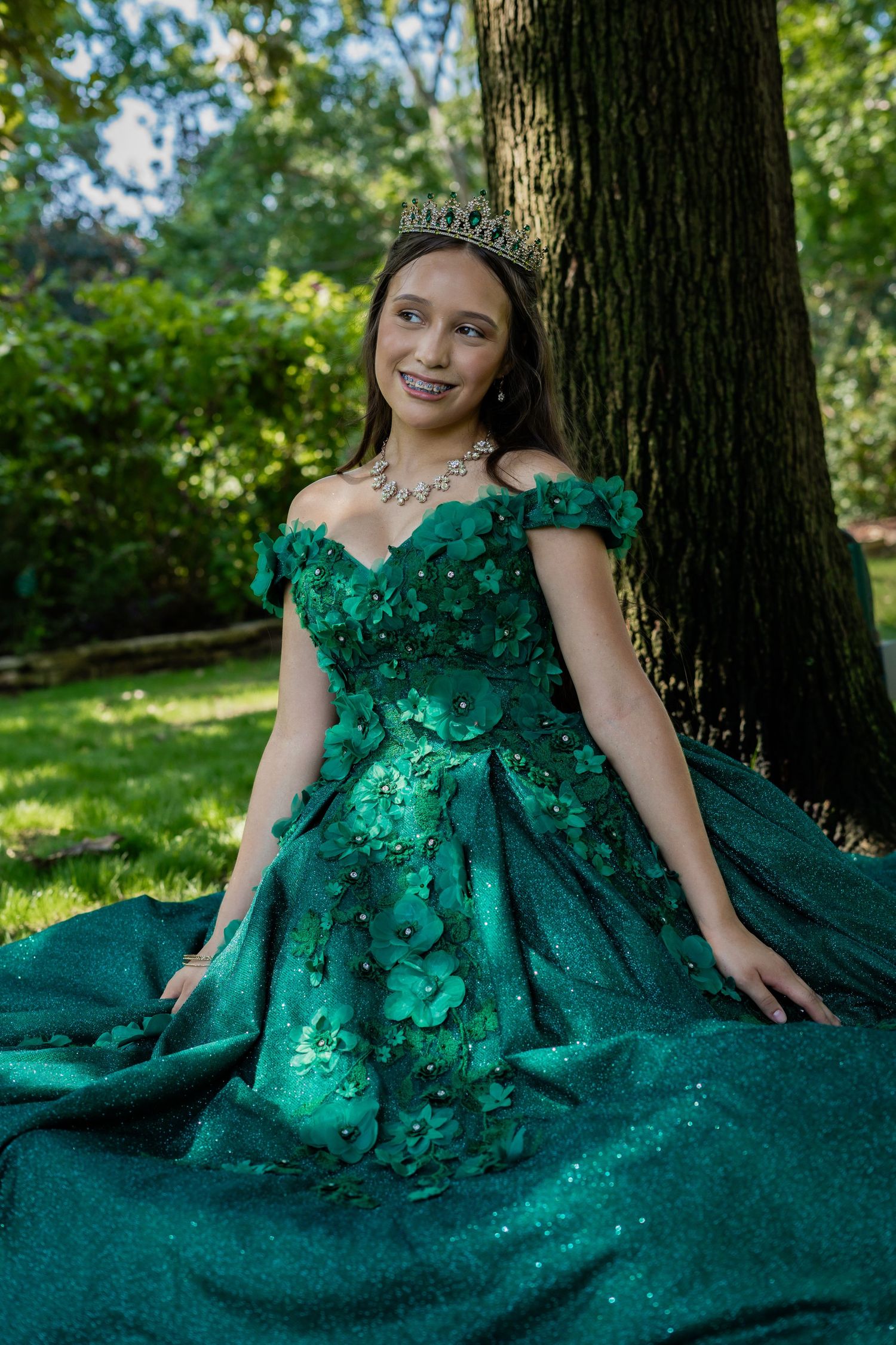 Young girl in emerald green quinceañera gown with floral details and tiara sits outdoors by a tree.