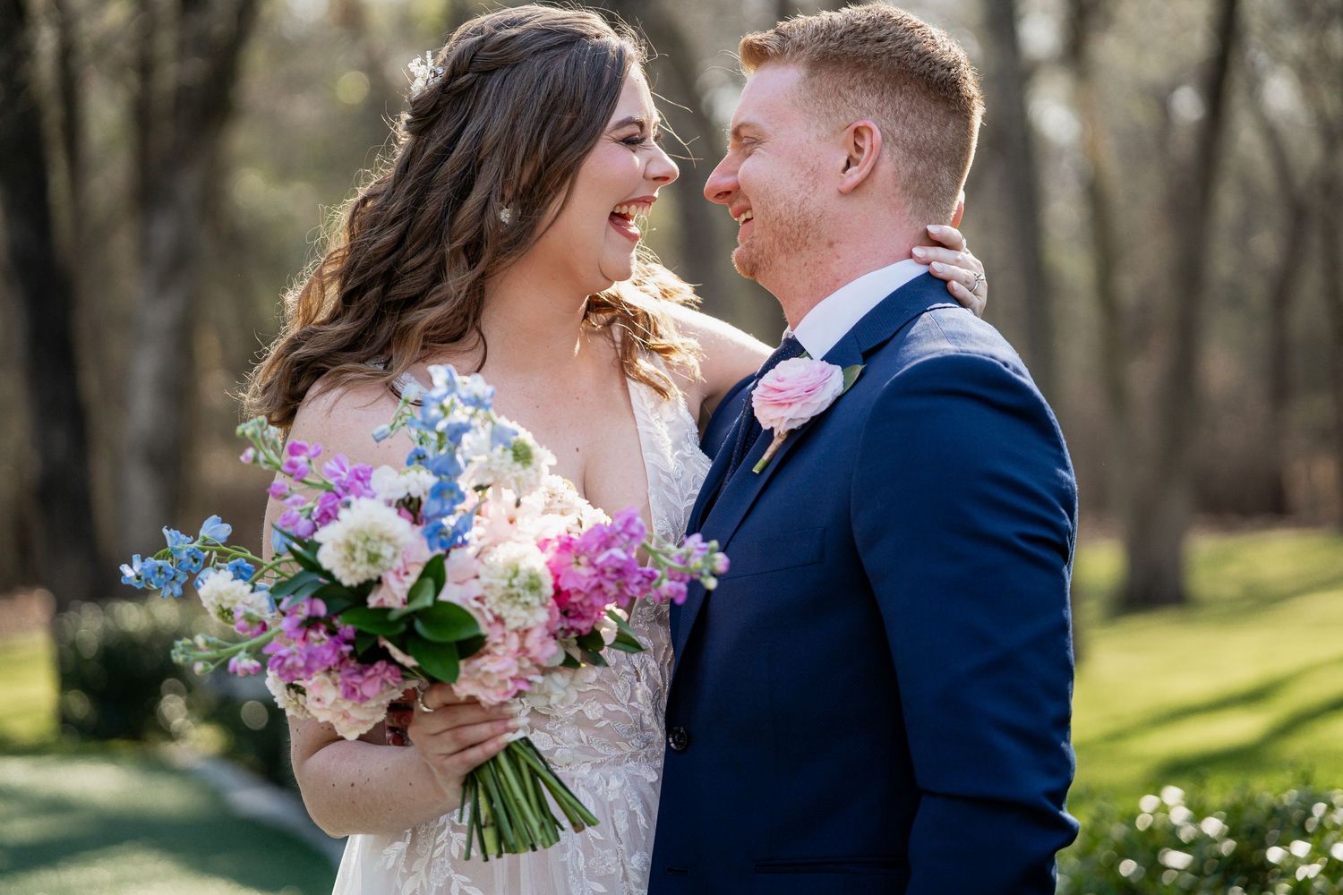 Bride and groom smiling at each other outdoors, bride holding colorful spring bouquet, groom in navy suit with pink boutonniere.