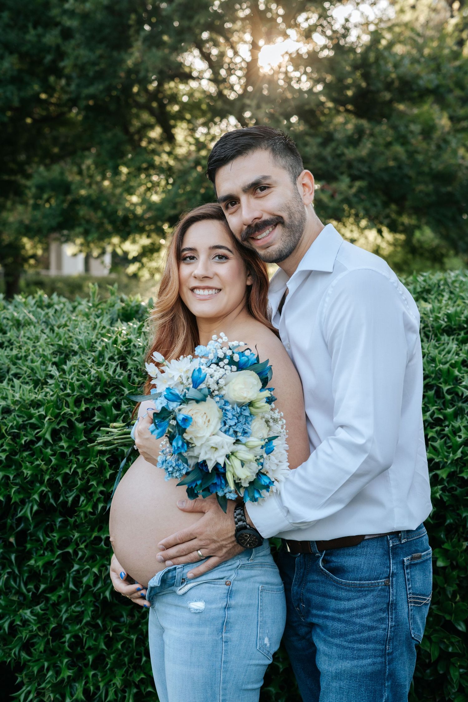 Smiling pregnant couple poses outdoors holding blue and white flower bouquet, man embracing woman's baby bump near green hedges.