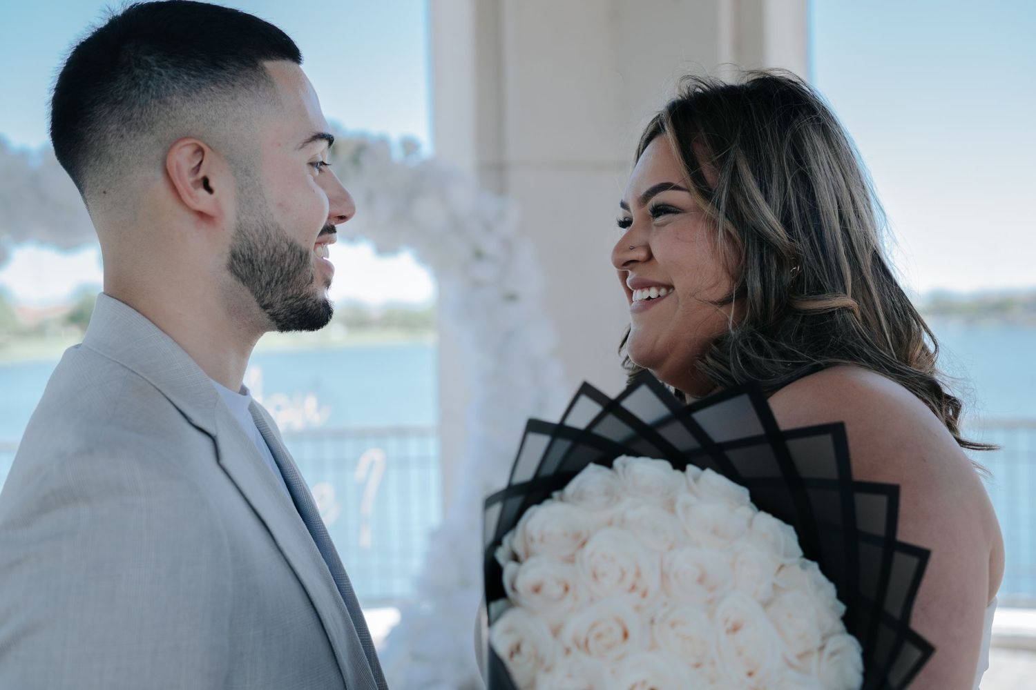 Couple sharing a romantic moment, man in gray suit, woman holding large white rose bouquet, waterfront venue backdrop.