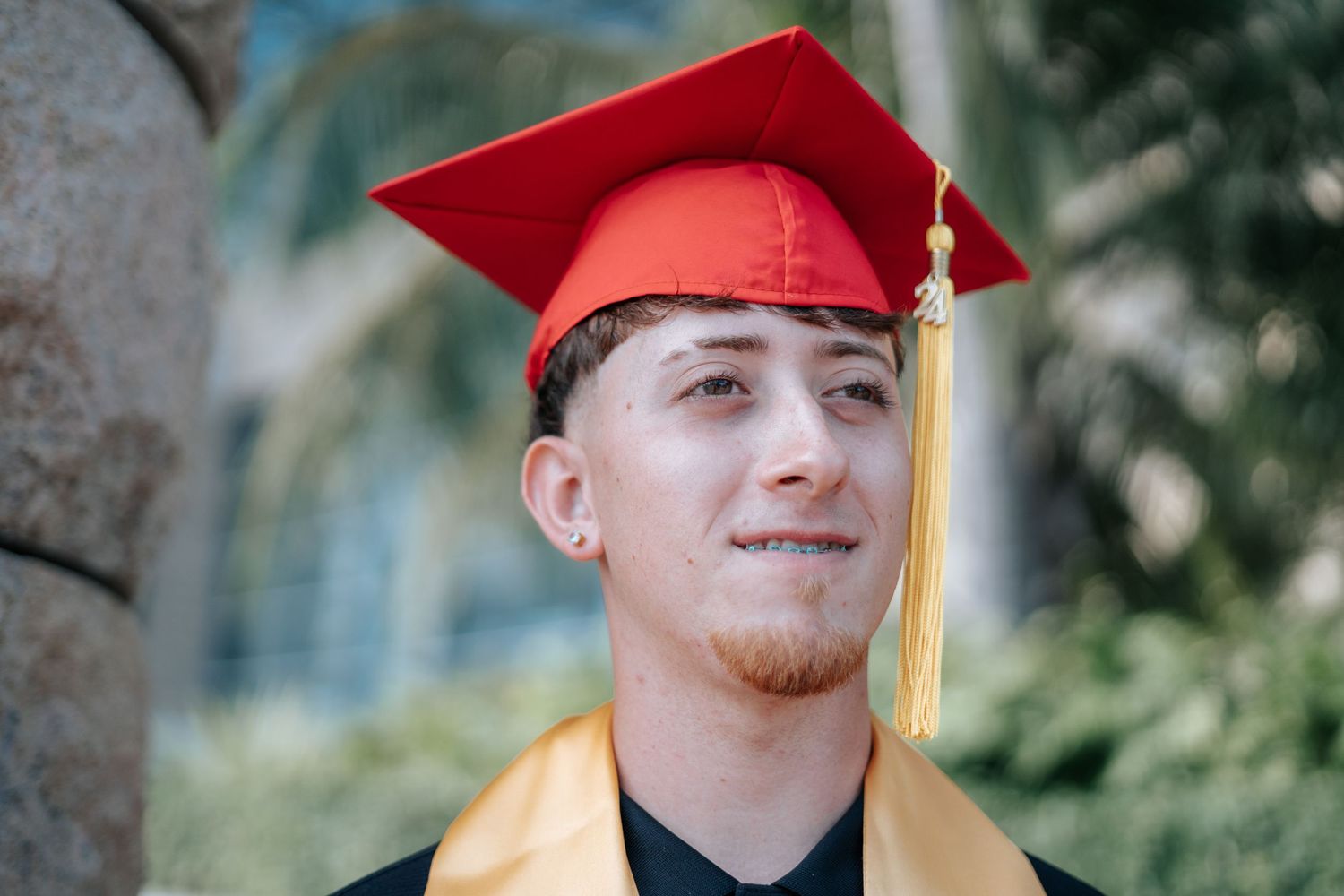 Young man smiling in red graduation cap and gold stole outdoors on graduation day.