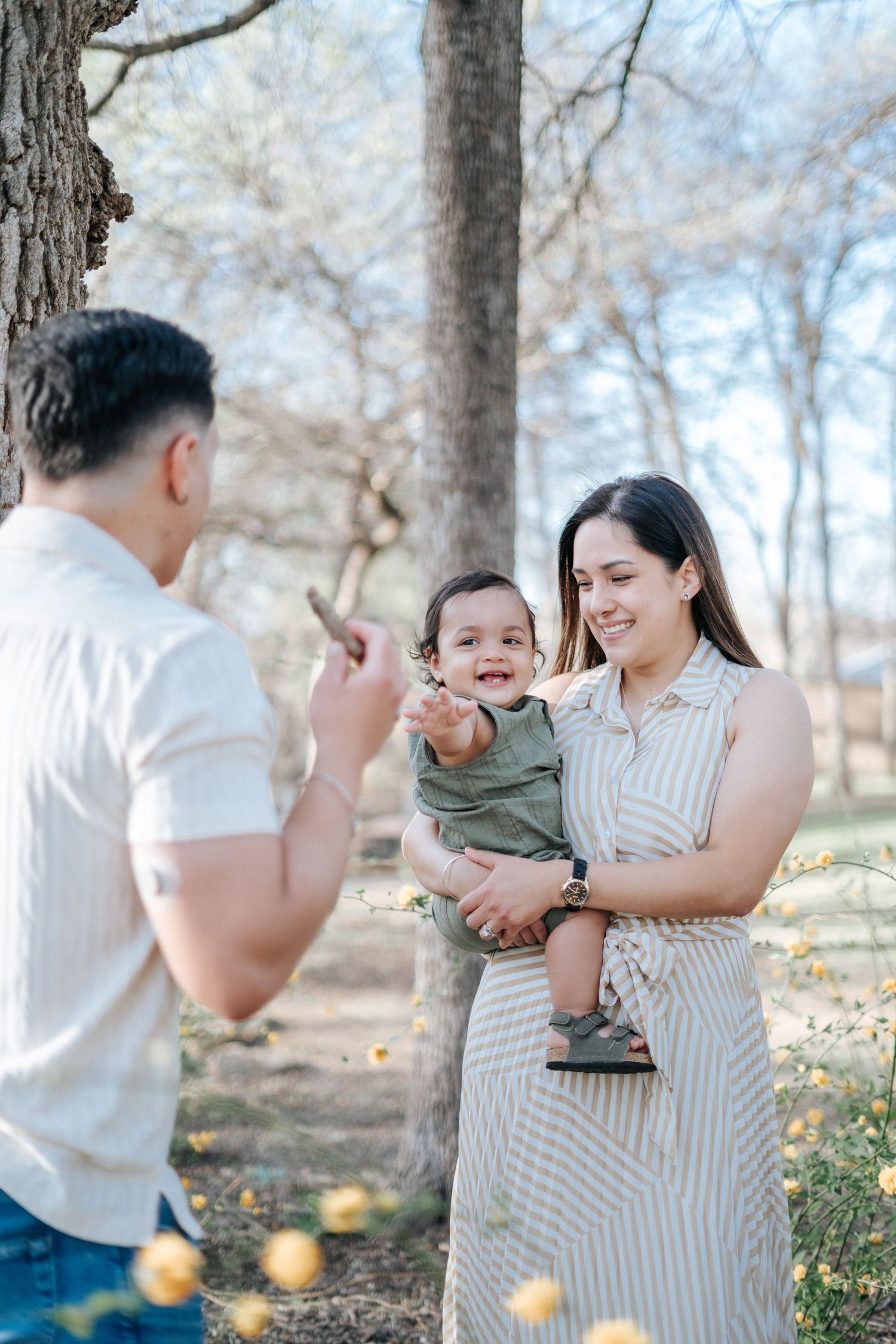 A young family enjoys a sunny day outdoors, with a smiling mother holding her baby boy near a tree.