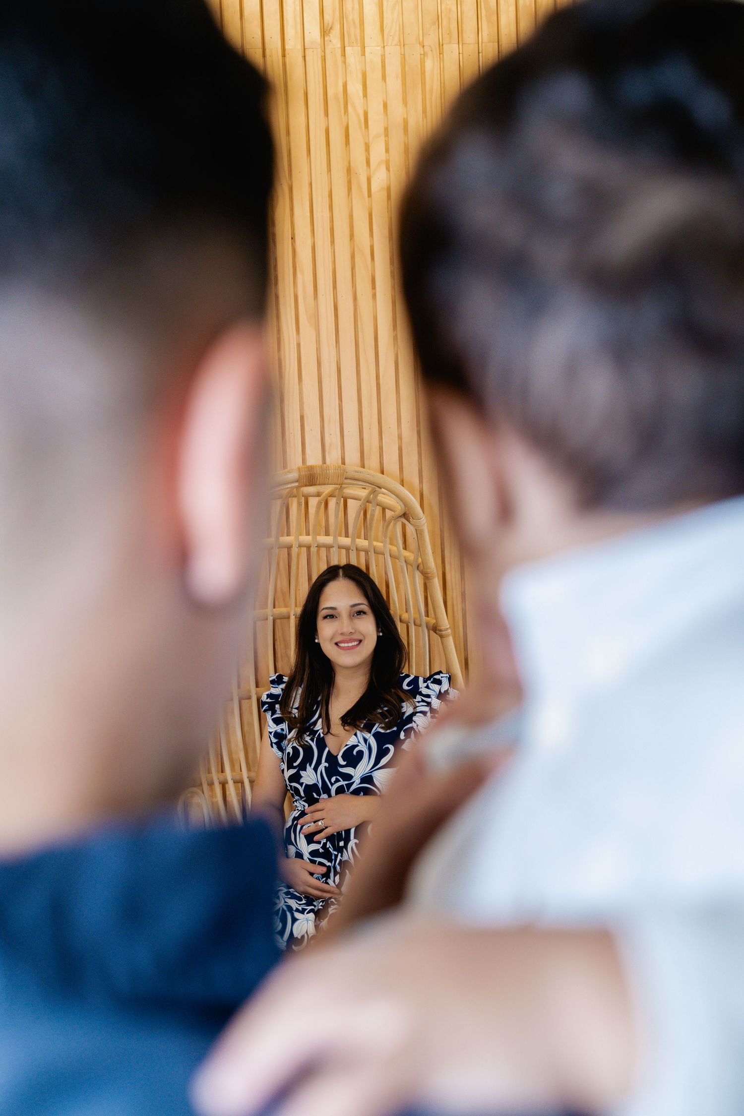 Woman in blue floral dress smiling, framed between two people in a rattan decorated interior setting.