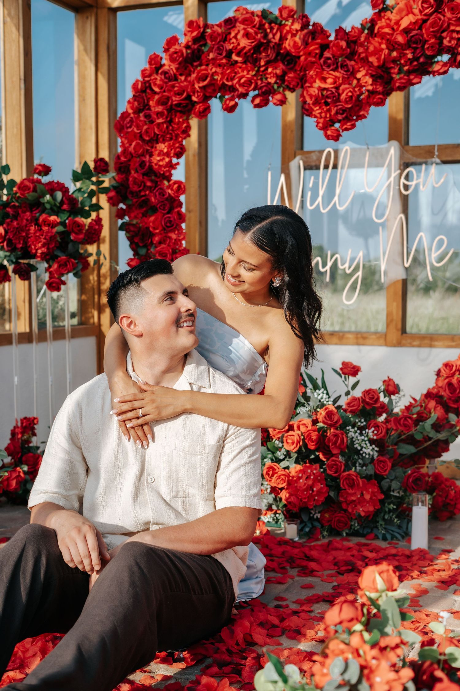 Romantic couple surrounded by red roses and a Will You Marry Me neon sign during a heartfelt marriage proposal.