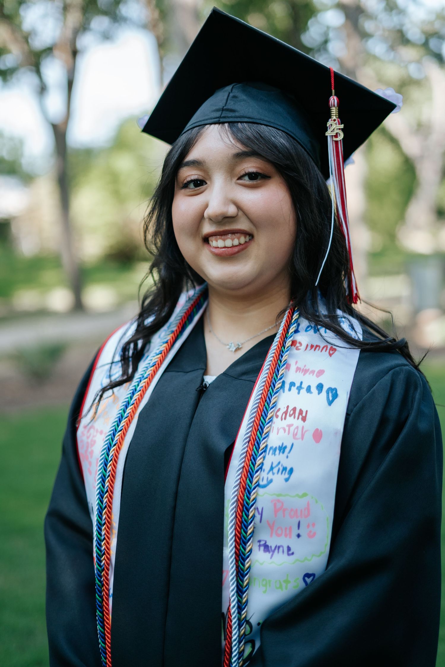 Smiling graduate in black cap and gown with decorated stole and colorful honor cords outdoors.