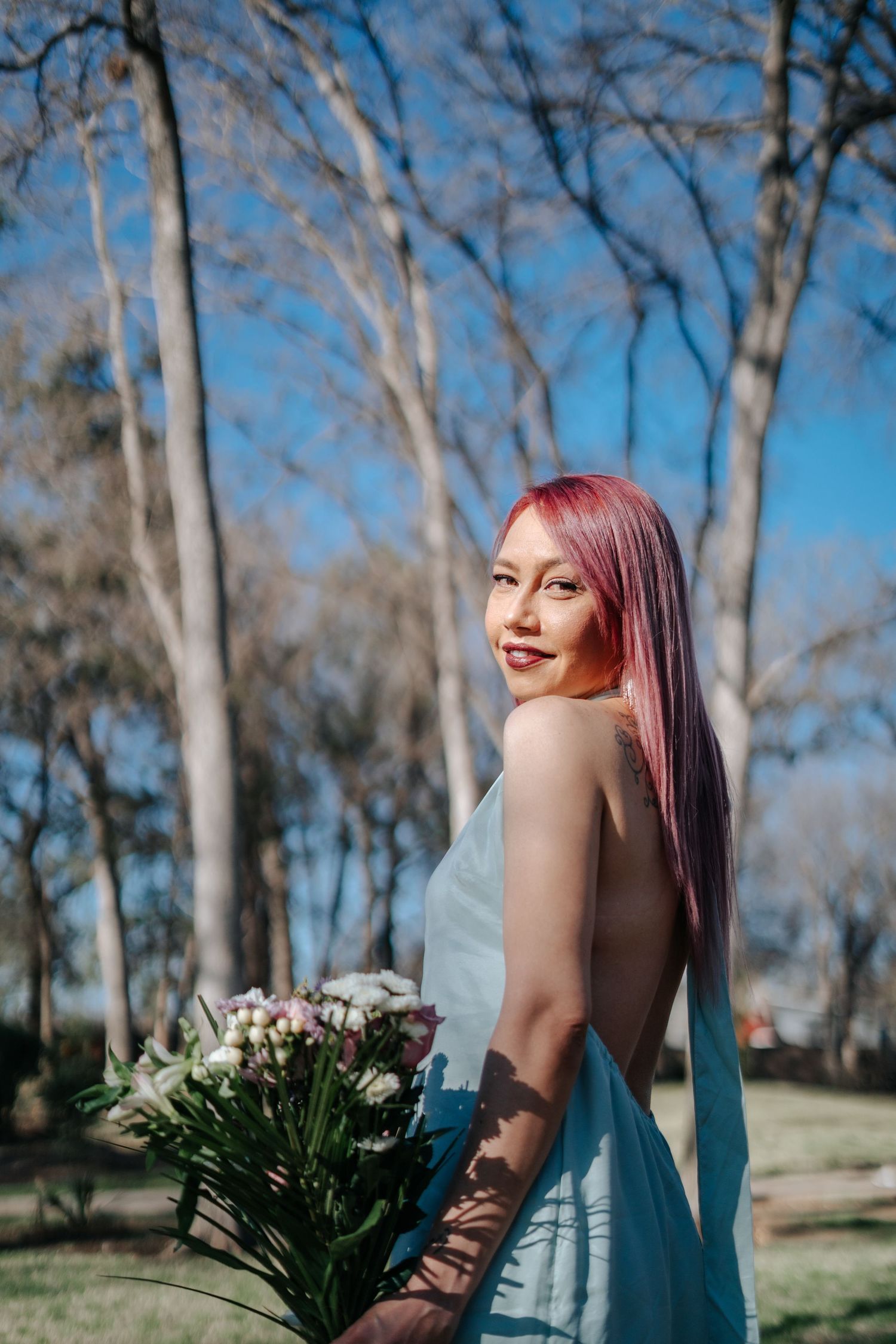 Young woman with pink hair in a light blue dress holding a bouquet of flowers outdoors among bare trees.