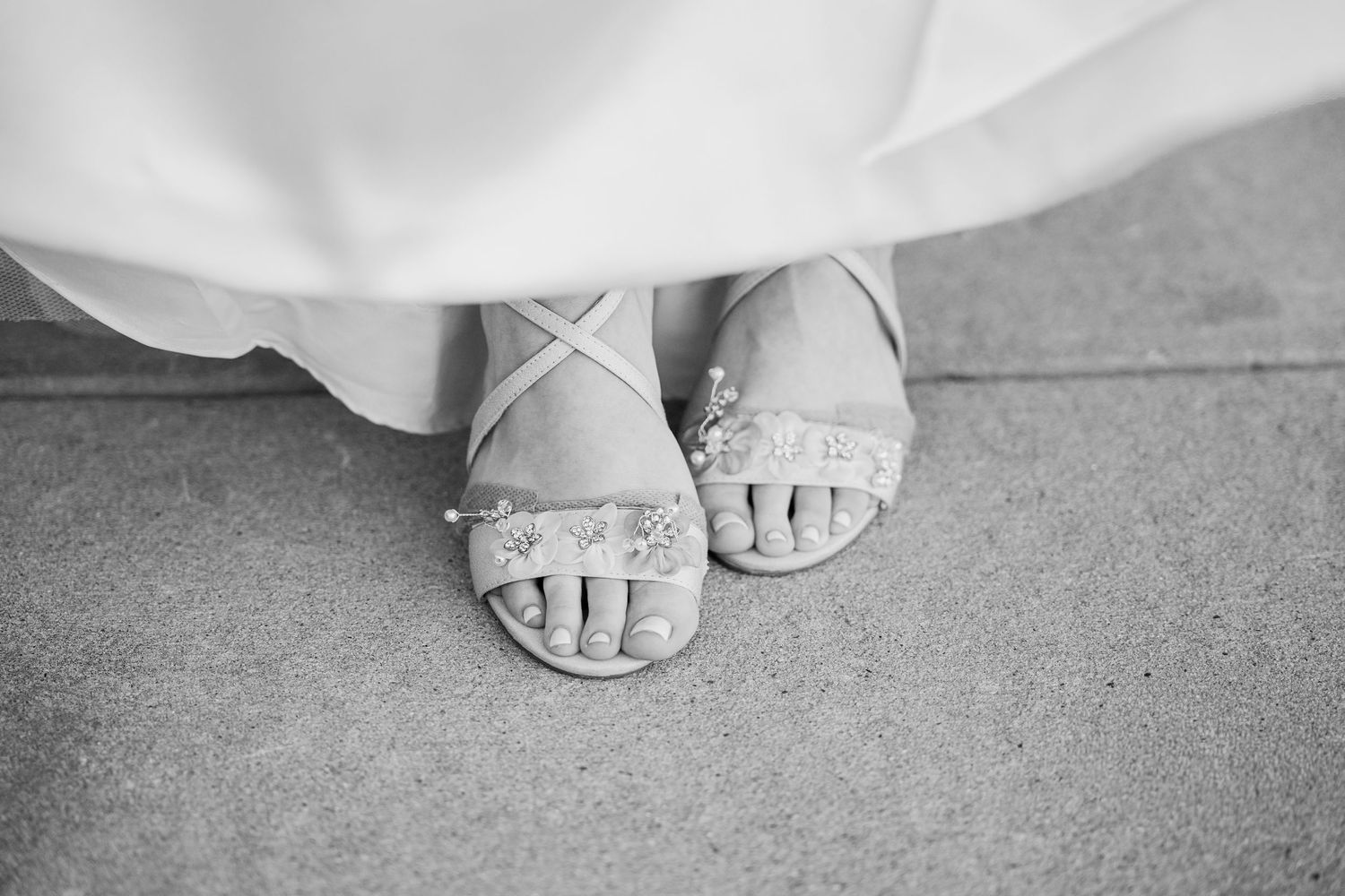 Black and white photo of a bride wearing elegant embellished strappy sandals beneath her white wedding gown.