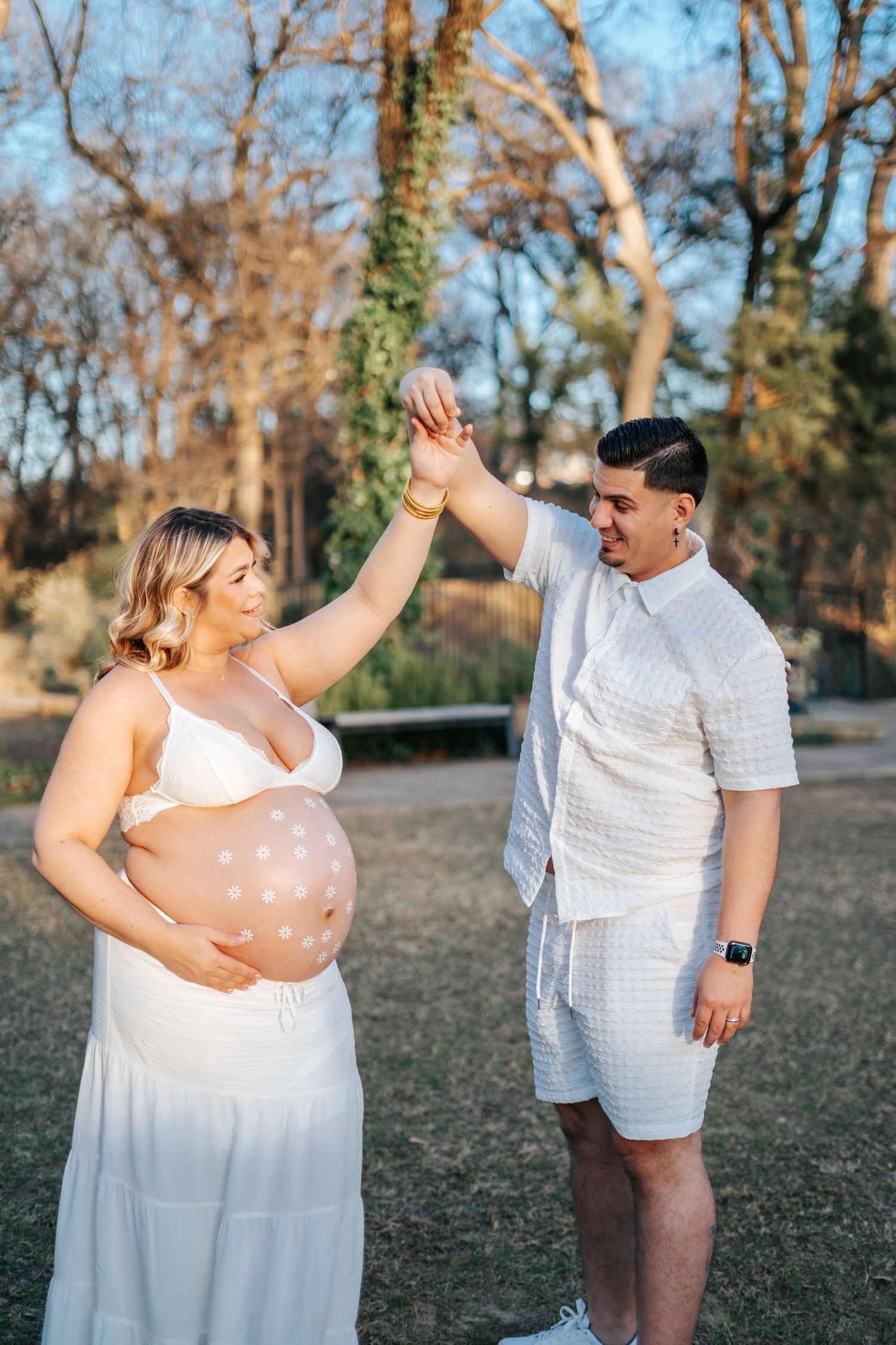 Pregnant woman in white dress holds belly while partner raises her hand in sunny park maternity photoshoot.