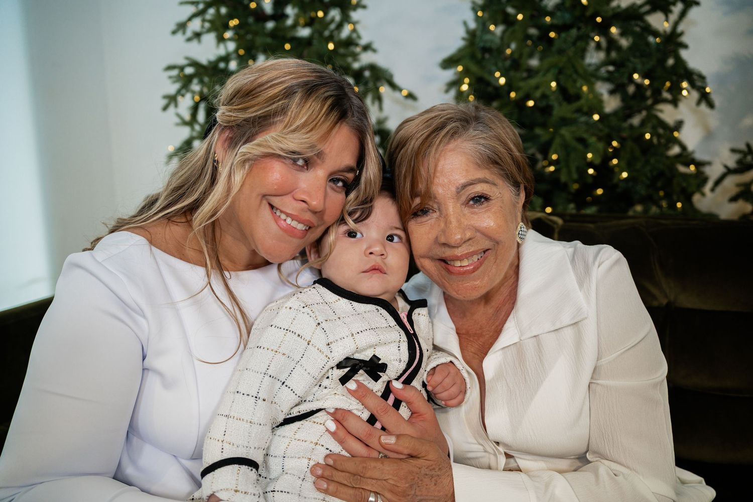 Three generations of women pose together near a Christmas tree, with a baby in the center wearing a festive outfit.