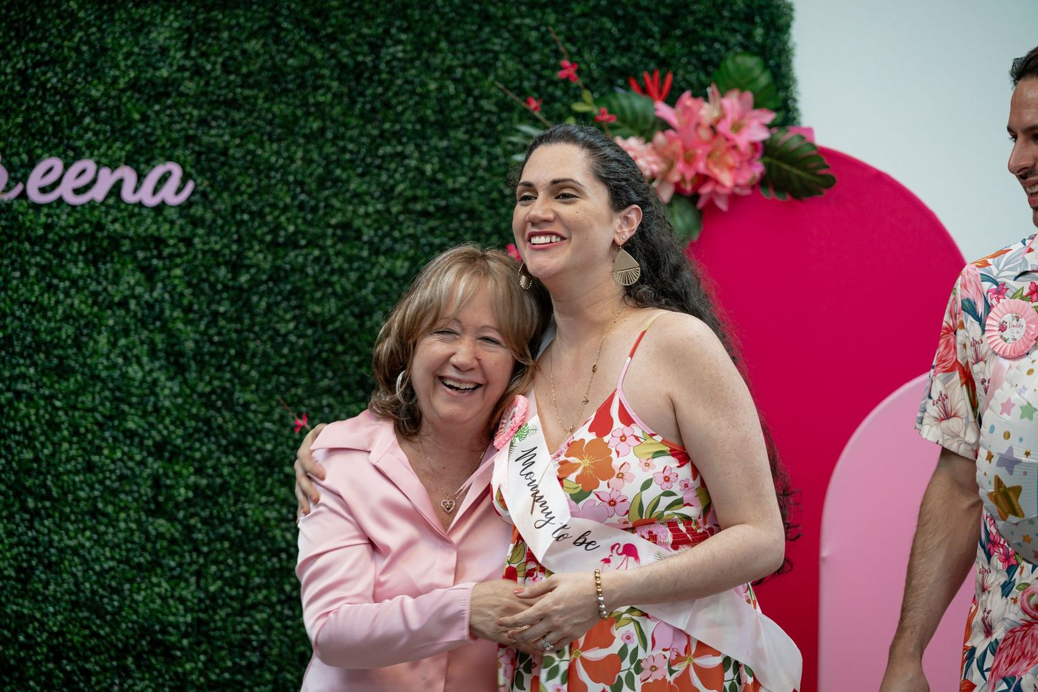 Two women smiling and hugging at a floral event, one wearing a sash, in front of a green wall backdrop.