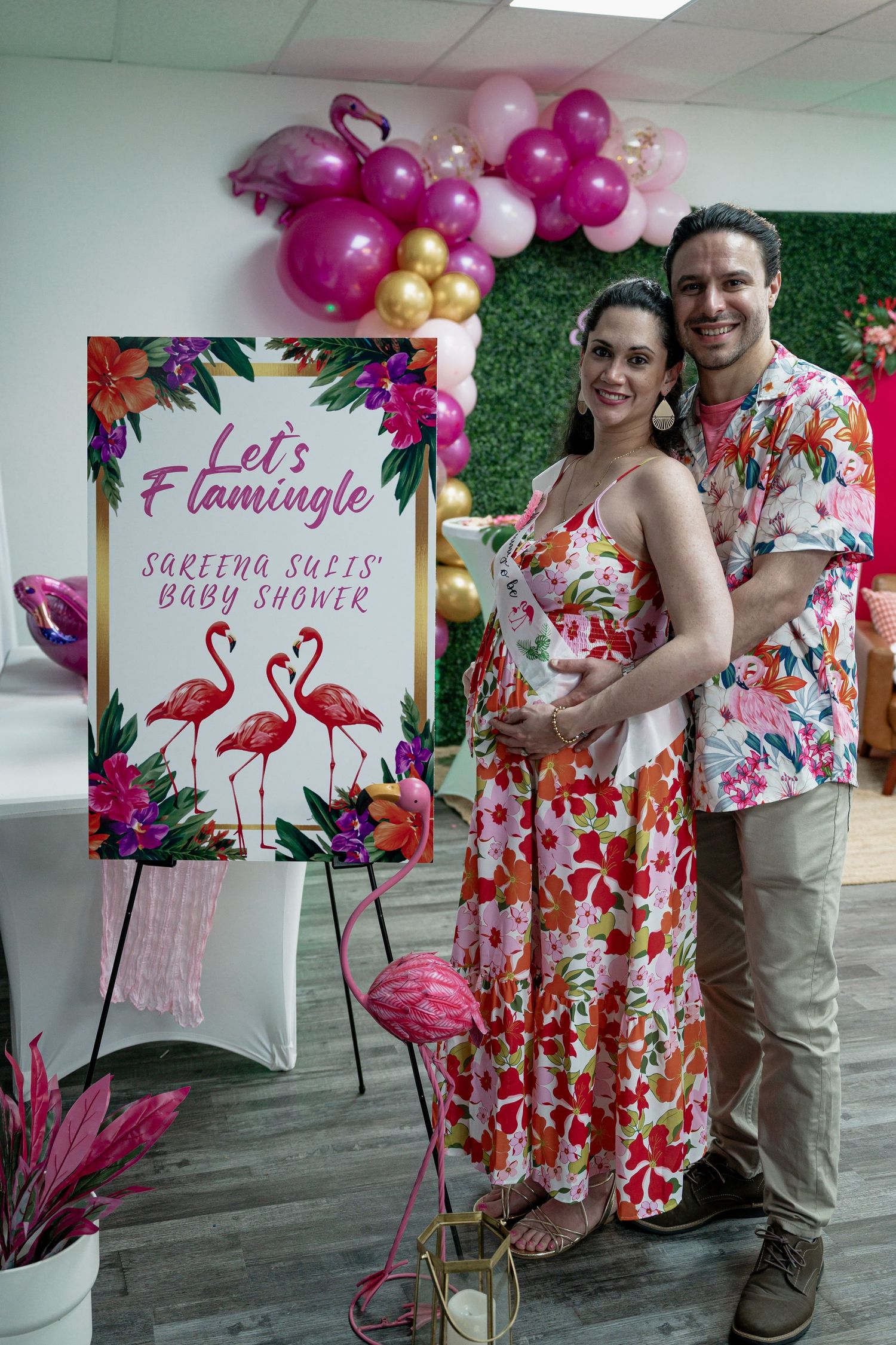Couple posing at a flamingo-themed baby shower with pink balloons and a colorful floral backdrop.
